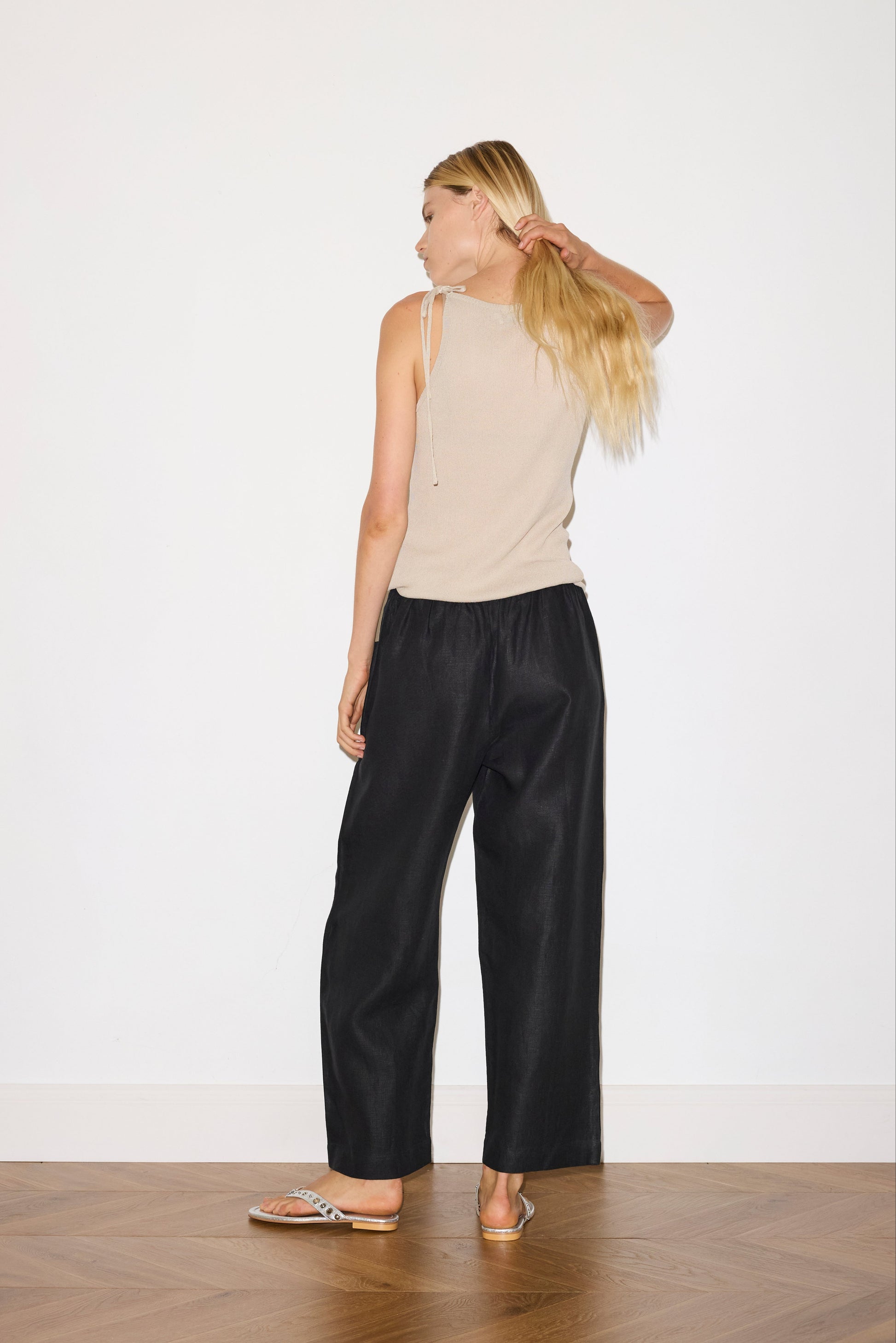 blonde haired female model wears black an ankle length, straight leg pant features a soft minimal elastic waist with front seam pockets and button detail, paired with a beige a minimalist tank-style top featuring raw edge detailing and an asymmetrical tie strap design for a unique and adjustable fit. made with a lightweight ribbed texture