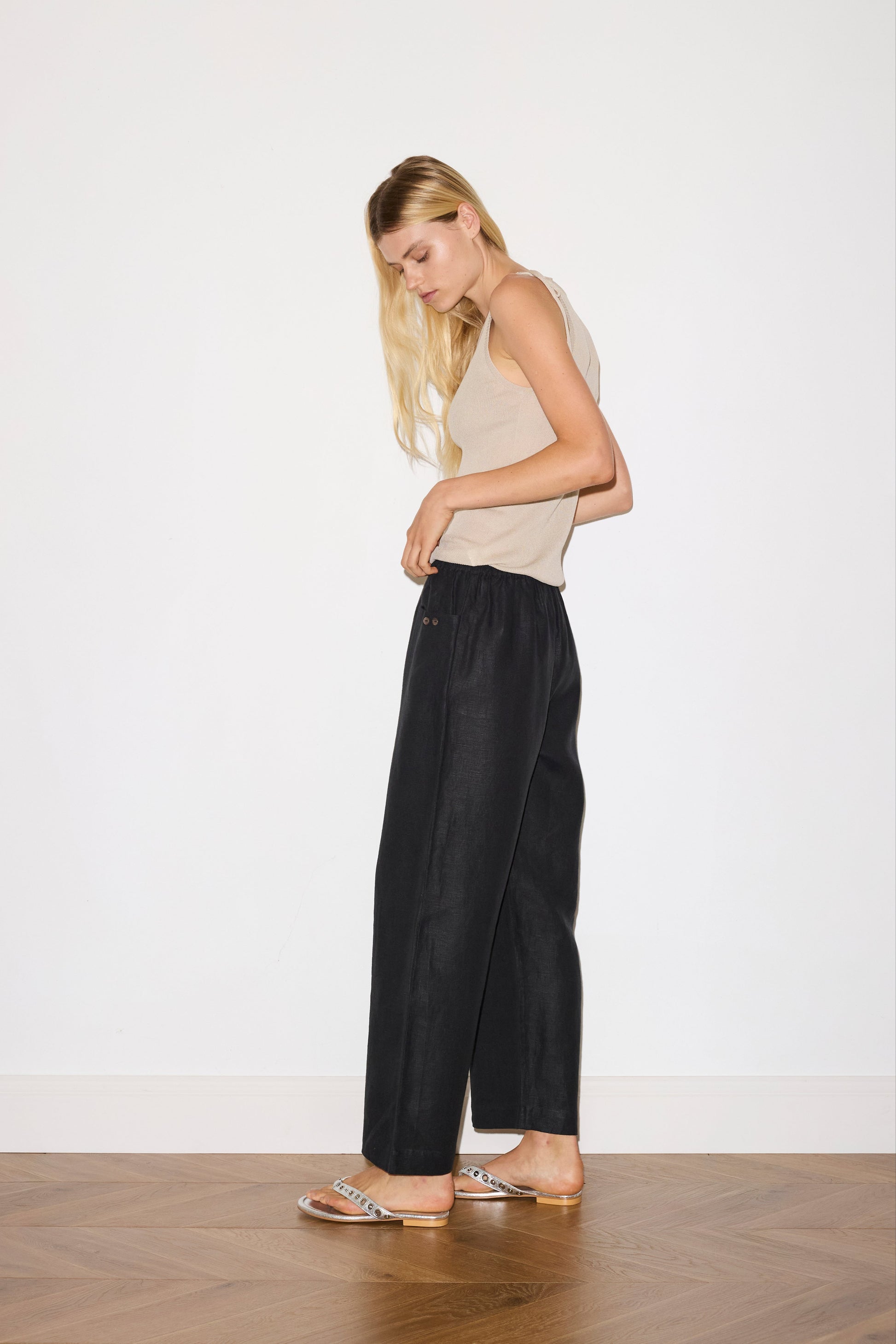 blonde haired female model wears black an ankle length, straight leg pant features a soft minimal elastic waist with front seam pockets and button detail, paired with a beige a minimalist tank-style top featuring raw edge detailing and an asymmetrical tie strap design for a unique and adjustable fit. made with a lightweight ribbed texture