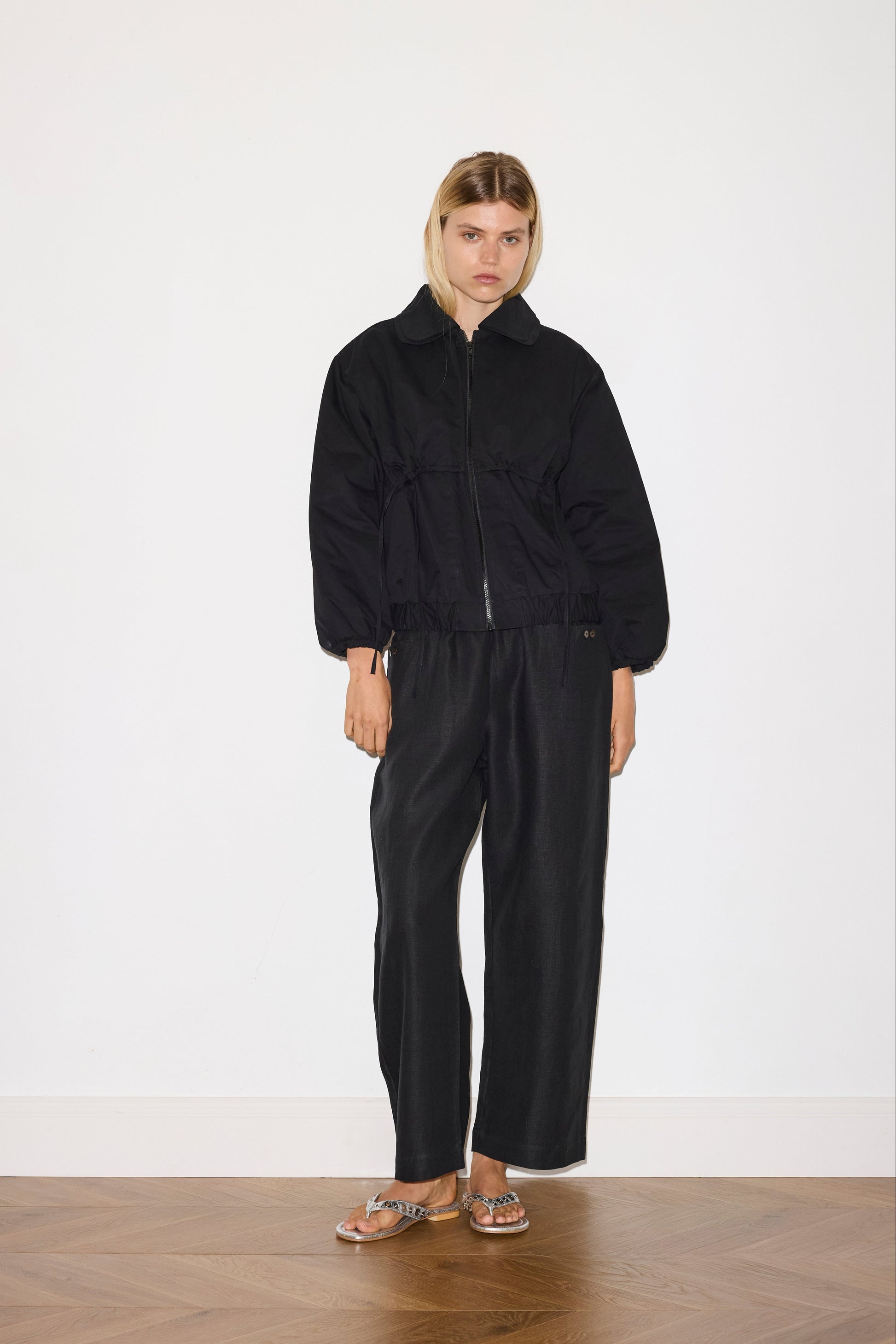 blonde haired female model wears black an ankle length, straight leg pant features a soft minimal elastic waist with front seam pockets and button detail, paired with a black organic cotton zip jacket 