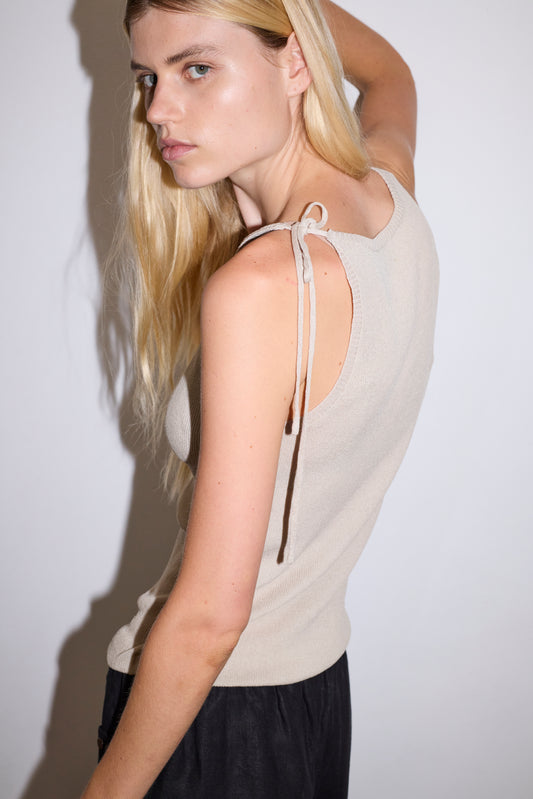 blonde female model wears a beige minimalist tank-style top featuring raw edge detailing and an asymmetrical tie strap design for a unique and adjustable fit. made with a lightweight ribbed texture, worn with a black ankle length, straight leg pant features a soft minimal elastic waist with front seam pockets and button detail