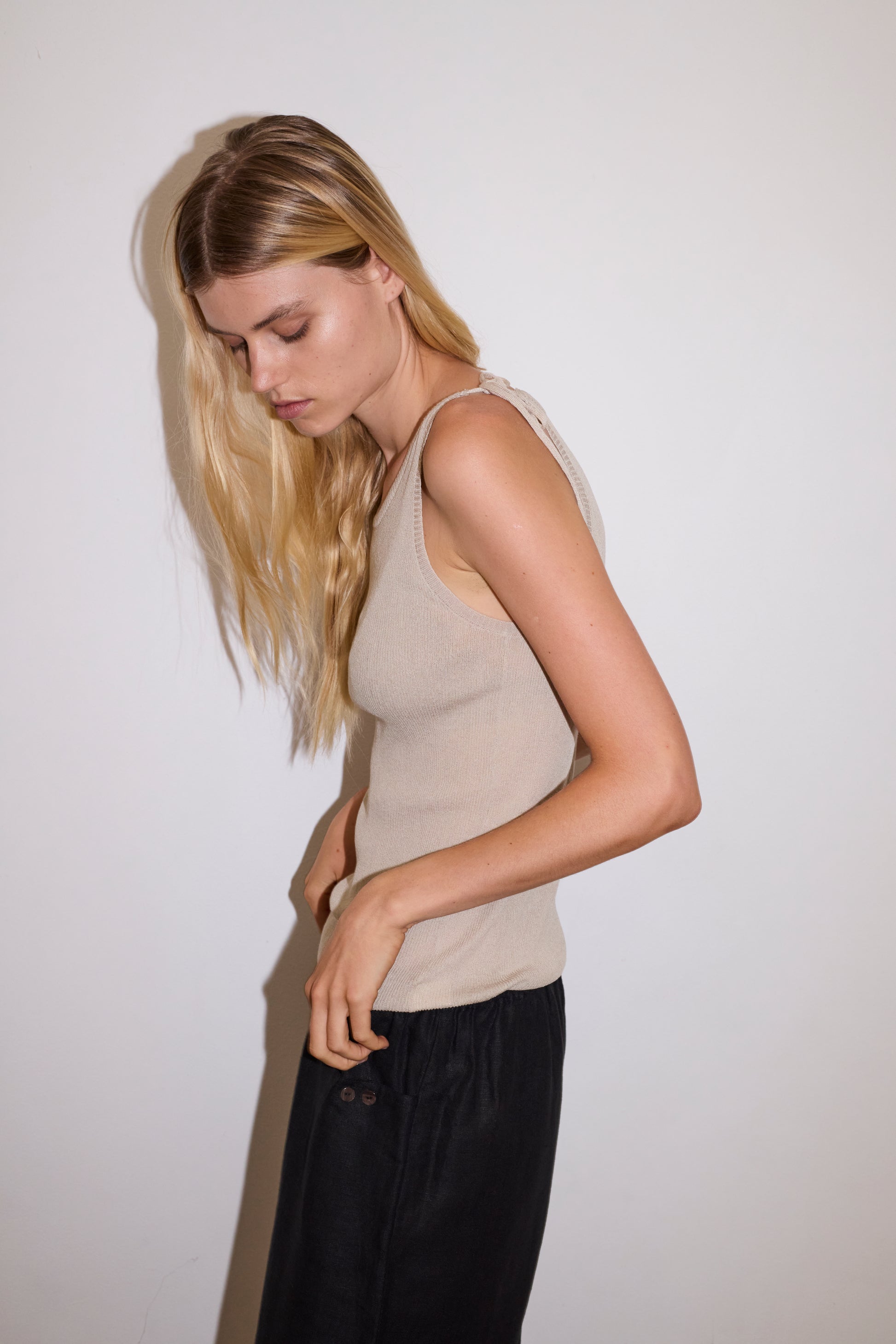 blonde female model wears a beige minimalist tank-style top featuring raw edge detailing and an asymmetrical tie strap design for a unique and adjustable fit. made with a lightweight ribbed texture, worn with a black ankle length, straight leg pant features a soft minimal elastic waist with front seam pockets and button detail