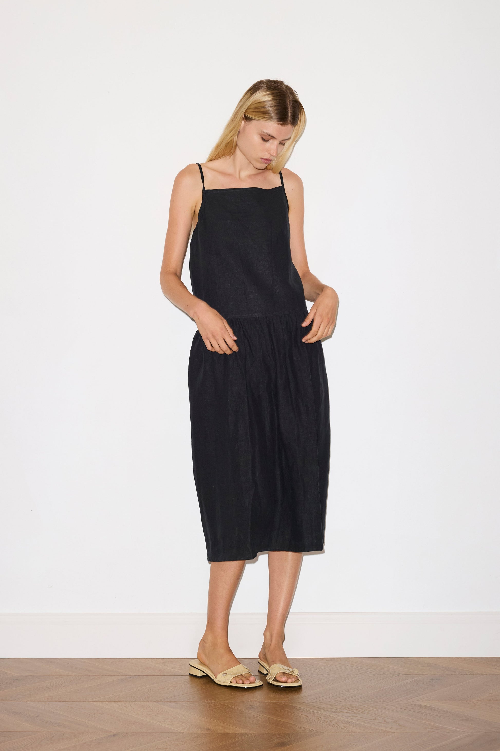 blonde haired female model wears a black relaxed mid-length dress featuring a drop waist with soft gathered skirt, gathering under the armholes and adjustable tie straps at back