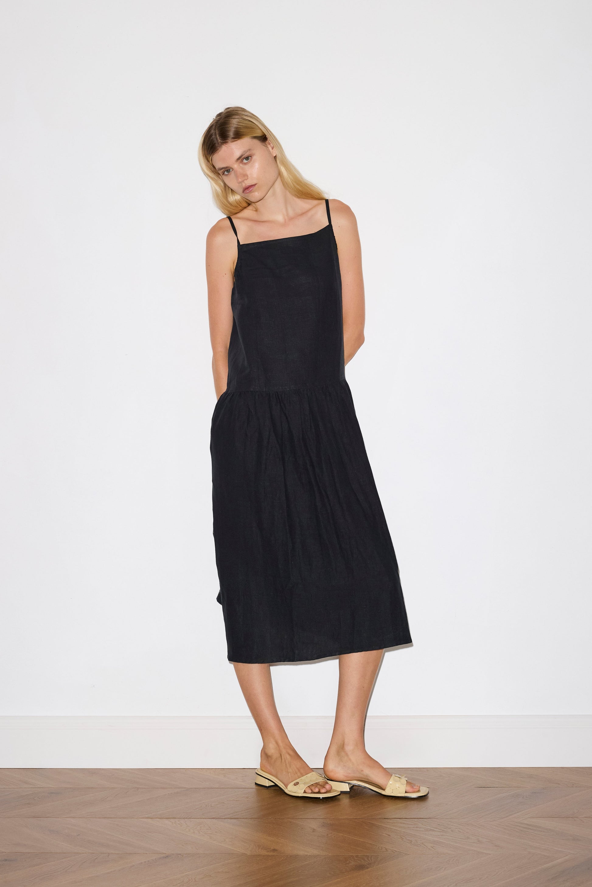 blonde haired female model wears a black relaxed mid-length dress featuring a drop waist with soft gathered skirt, gathering under the armholes and adjustable tie straps at back