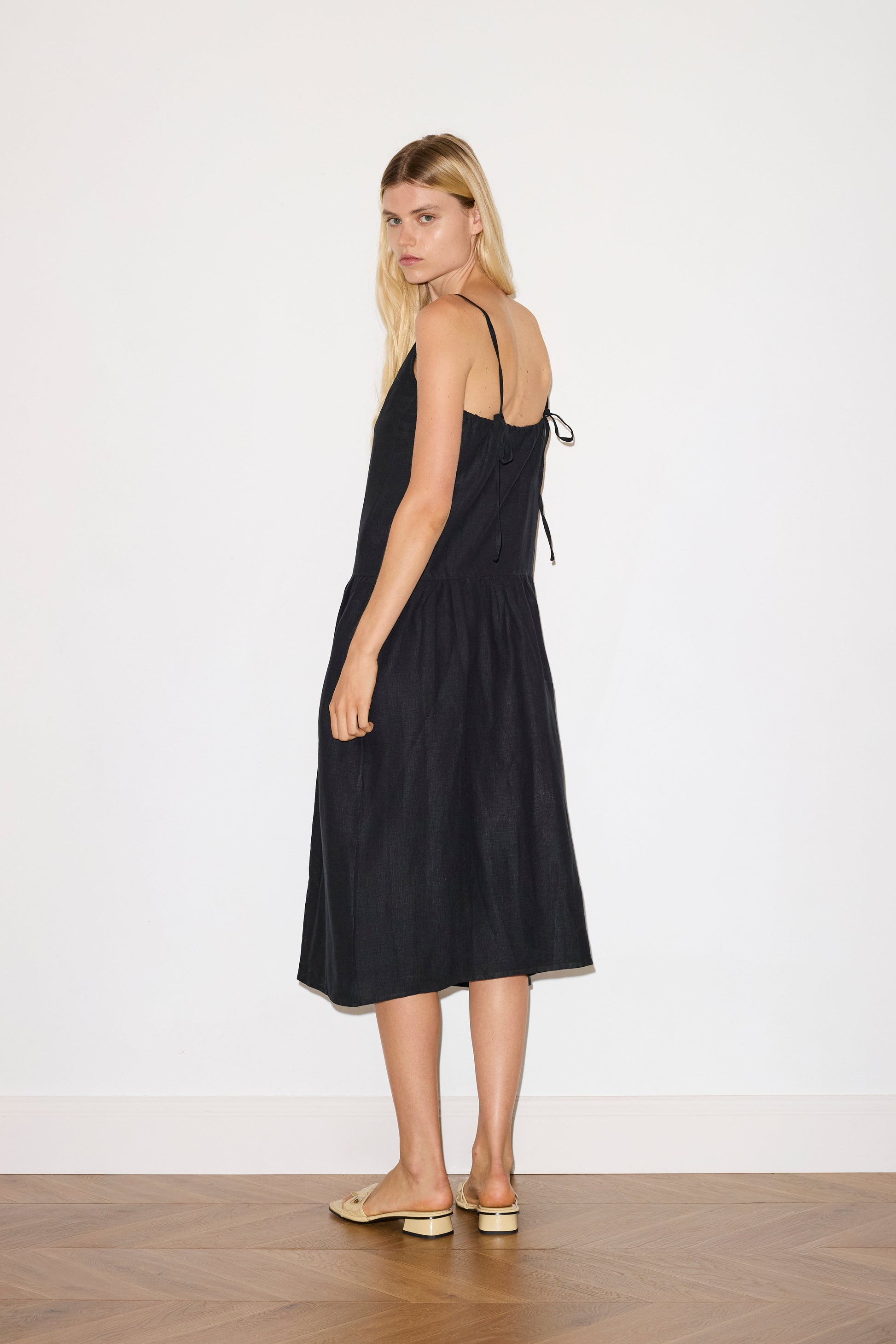 blonde haired female model wears a black relaxed mid-length dress featuring a drop waist with soft gathered skirt, gathering under the armholes and adjustable tie straps at back