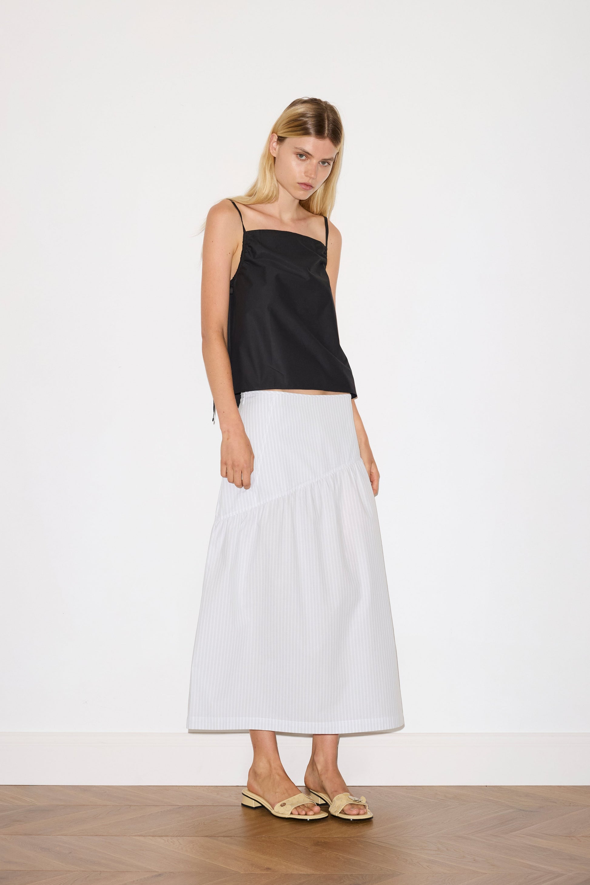 blonde female model wears a sage and white striped full-length skirt features single asymmetric design line on the front and back, a bound waistline for a structured fit, and a clean rolled hem, paired with a black a lightweight top featuring fixed gathered armholes and right hand side armhole keyhole and ties