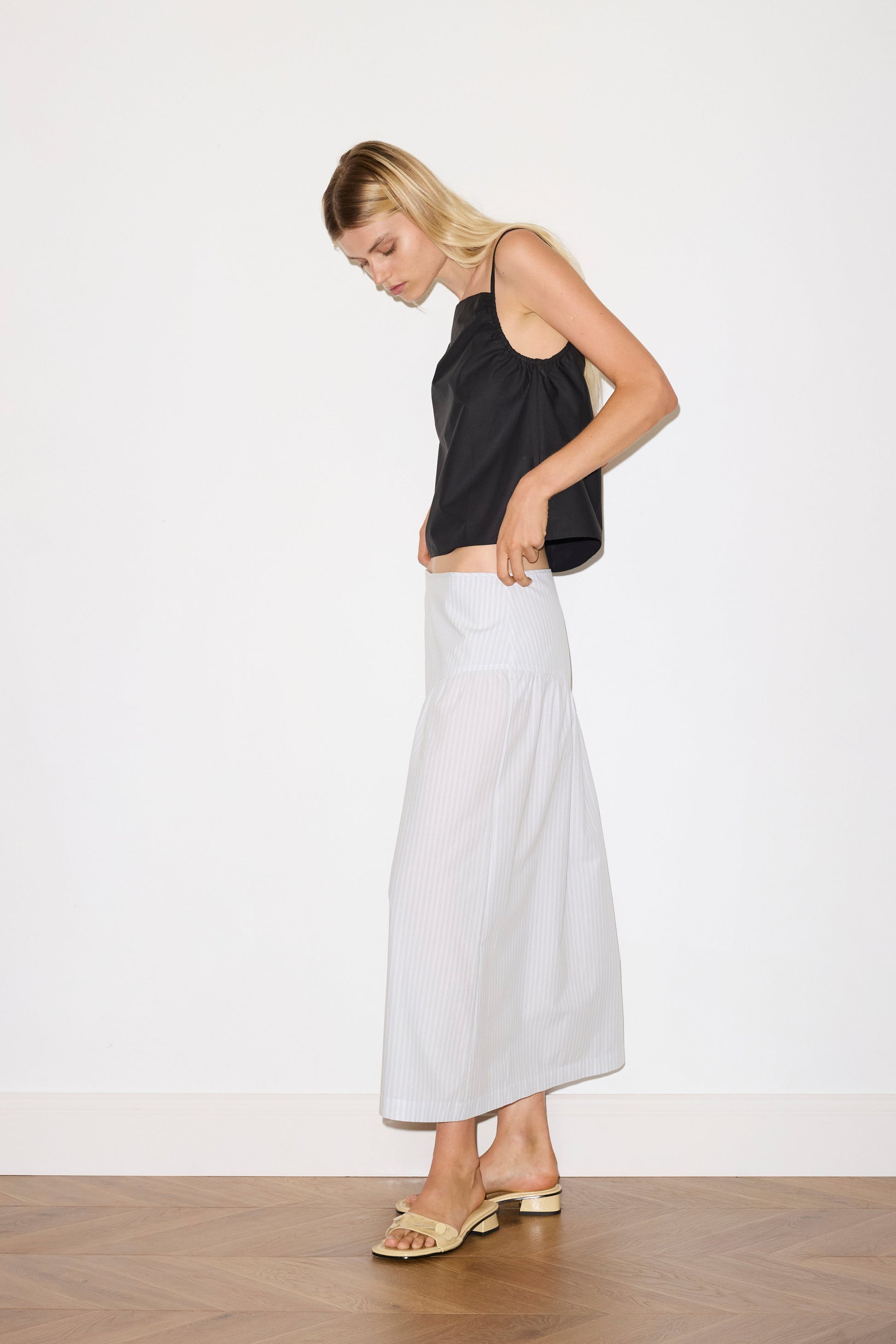 blonde female model wears a sage and white striped full-length skirt features single asymmetric design line on the front and back, a bound waistline for a structured fit, and a clean rolled hem, paired with a black a lightweight top featuring fixed gathered armholes and right hand side armhole keyhole and ties