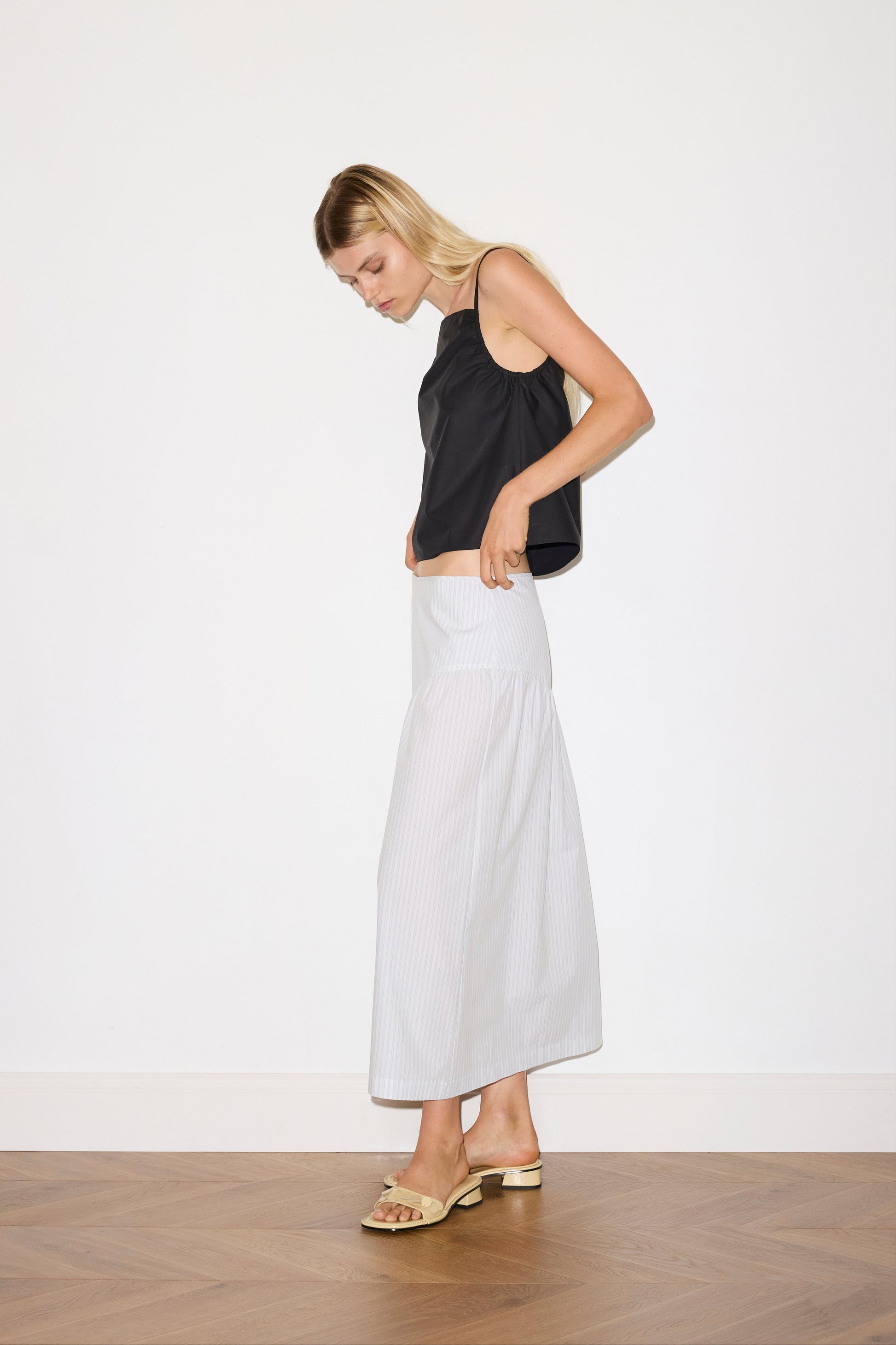 blonde female model wears a sage and white striped full-length skirt features single asymmetric design line on the front and back, a bound waistline for a structured fit, and a clean rolled hem, paired with a black a lightweight top featuring fixed gathered armholes and right hand side armhole keyhole and ties