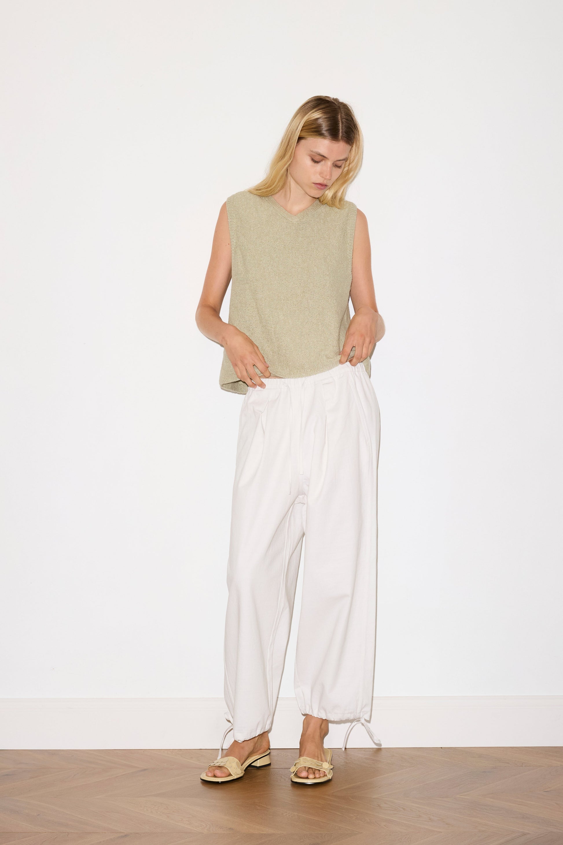 blond haired female model wears a sage lightweight, soft knit vest features a soft v neckline designed to be worn on its own or for light layering, paired with a white structured cotton twill trouser featuring a drawcord hem and waist and deep side pockets designed to sit mid-low waist for a relaxed look