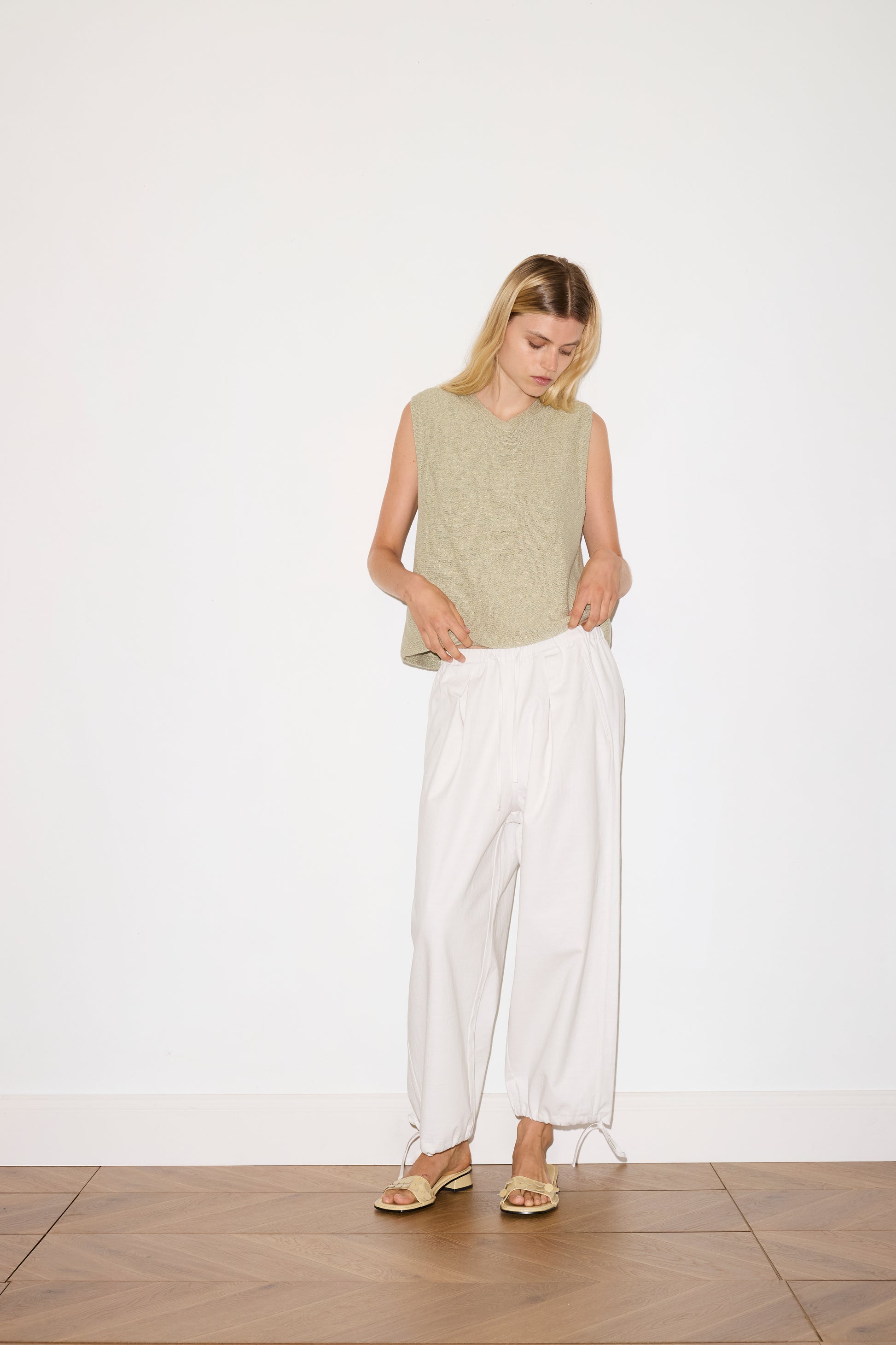 A woman stands indoors on a wooden floor, wearing a sleeveless beige top, the Drawcord Wide Leg Pant - White by Deiji Studios, and gold sandals, looking down with her hands in her pockets against a plain white wall.
