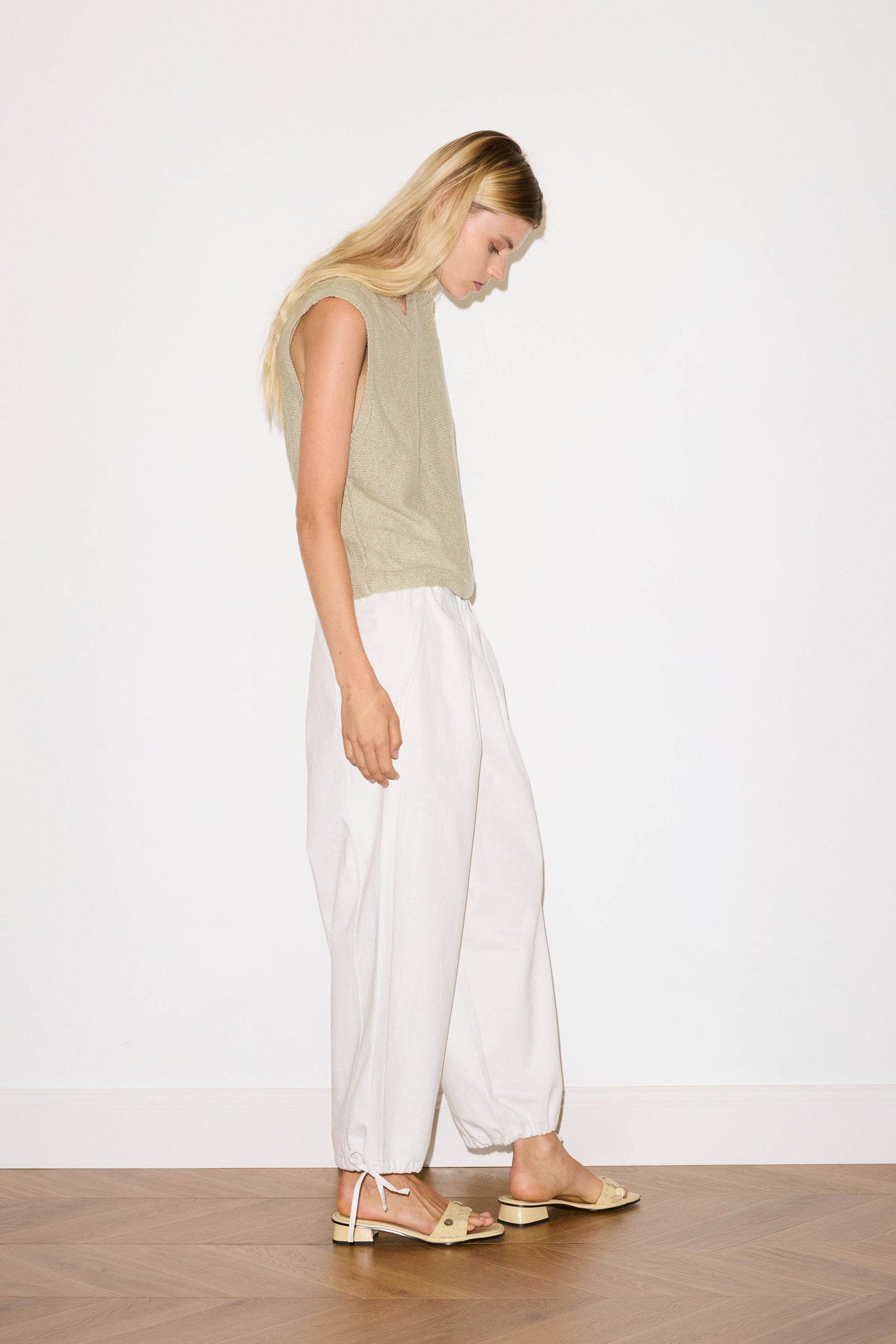 blond haired female model wears a sage lightweight, soft knit vest features a soft v neckline designed to be worn on its own or for light layering, paired with a white structured cotton twill trouser featuring a drawcord hem and waist and deep side pockets designed to sit mid-low waist for a relaxed look