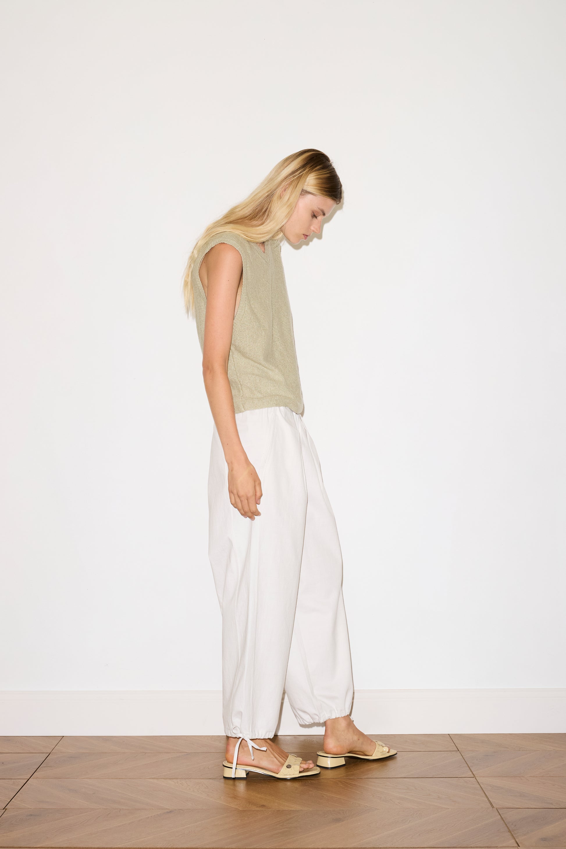 A woman with long blonde hair stands sideways on a wooden floor, looking down. She wears a sleeveless beige top, Drawcord Wide Leg Pant - White by Deiji Studios, and beige sandals against a plain white background.