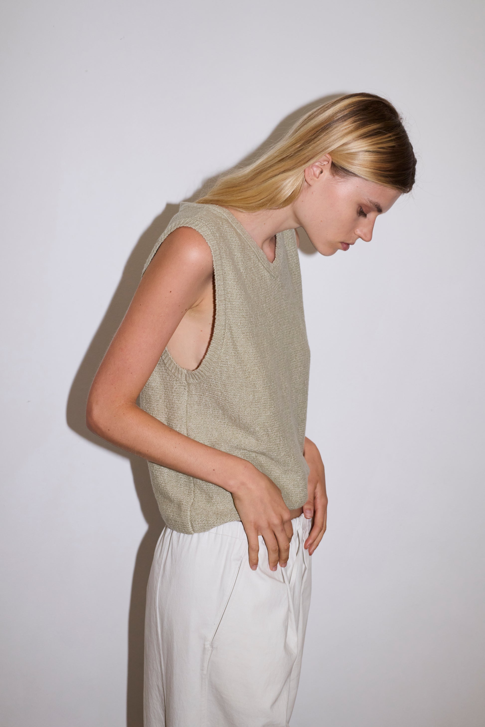 blond haired female model wears a sage lightweight, soft knit vest features a soft v neckline designed to be worn on its own or for light layering, paired with a white structured cotton twill trouser featuring a drawcord hem and waist and deep side pockets designed to sit mid-low waist for a relaxed look