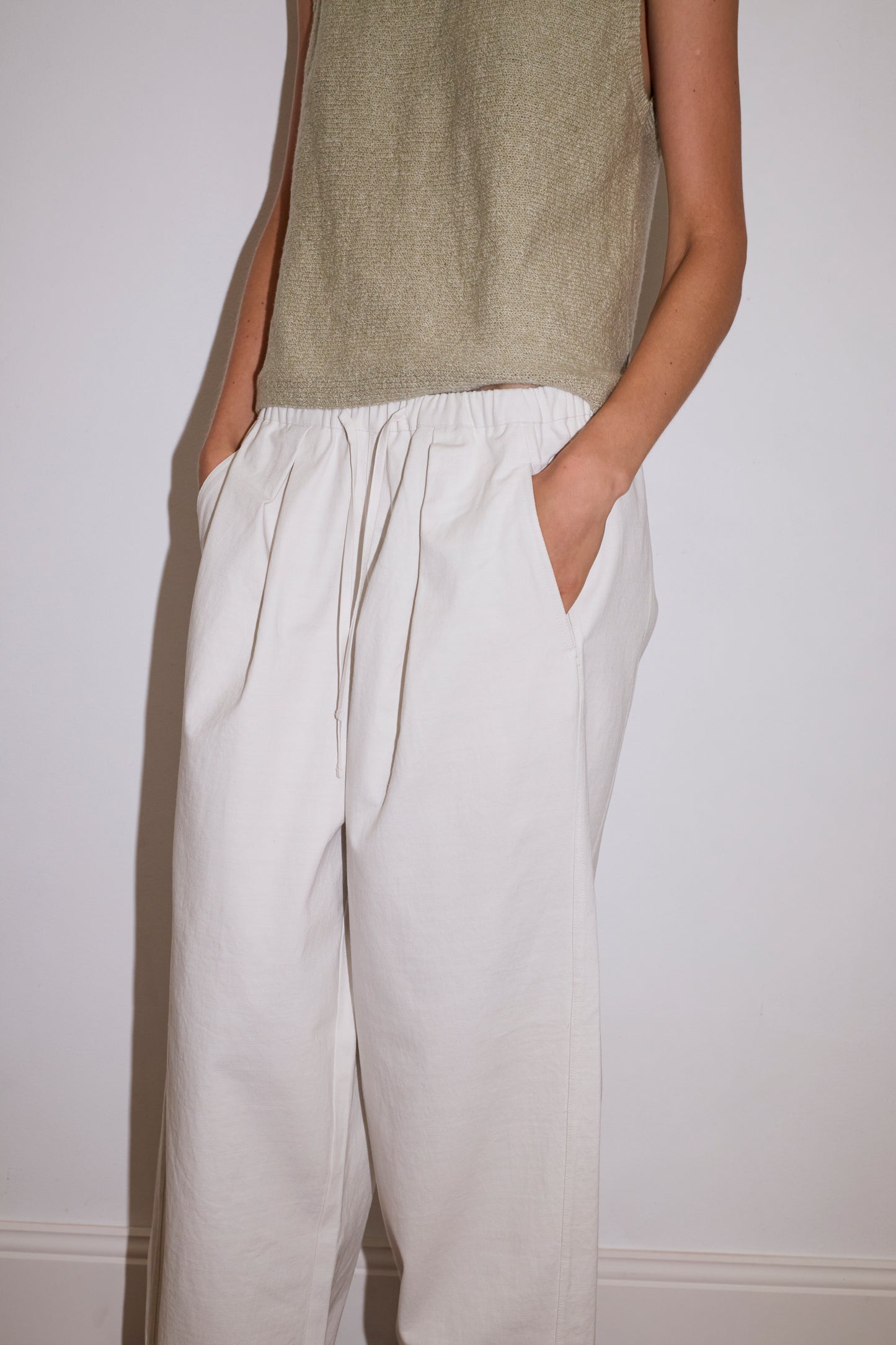 A person wears a sleeveless light olive top and the Drawcord Wide Leg Pant - White from Deiji Studios, made in organic cotton, standing against a plain white wall with hands in pockets.