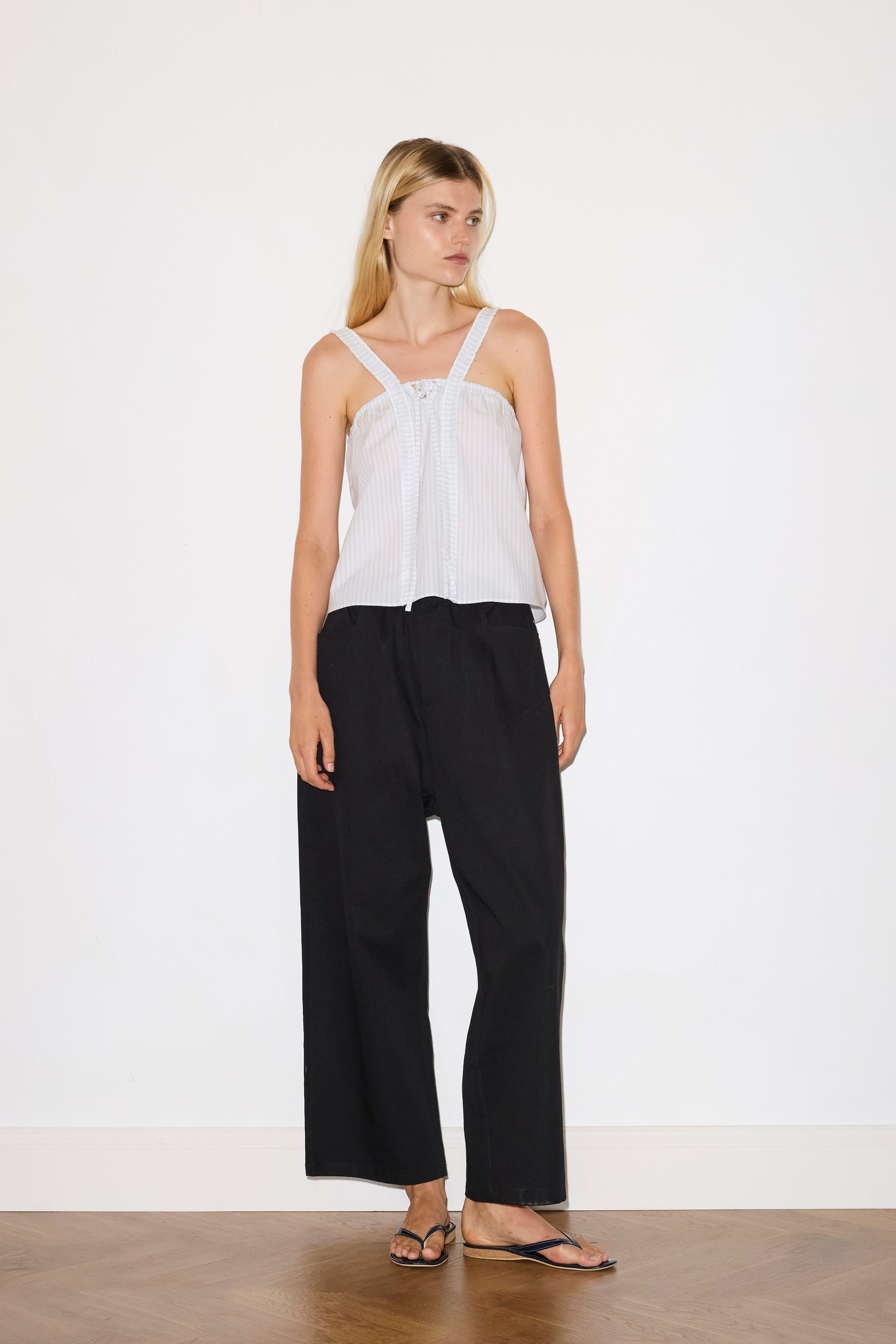 blonde female model wears a sage and white striped relaxed-fit top featuring self-fabric ruffles along the straps and center front, with a tie front detail. designed with elasticated gathering for ease and comfort, paired with a black Heavy- weighted relaxed-fit pant featuring a wide-leg silhouette, elasticated waistband, and square pocket detail