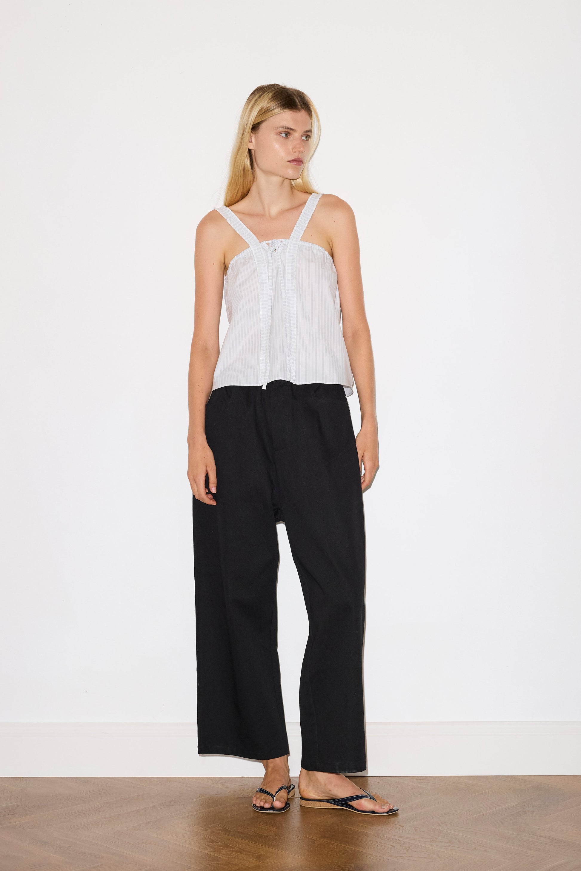 blonde haired female model wears a black Heavy- weighted relaxed-fit pant featuring a wide-leg silhouette, elasticated waistband, and square pocket detail, worn with a sage and white striped a relaxed-fit top featuring self-fabric ruffles along the straps and center front, with a tie front detail. designed with elasticated gathering for ease and comfort