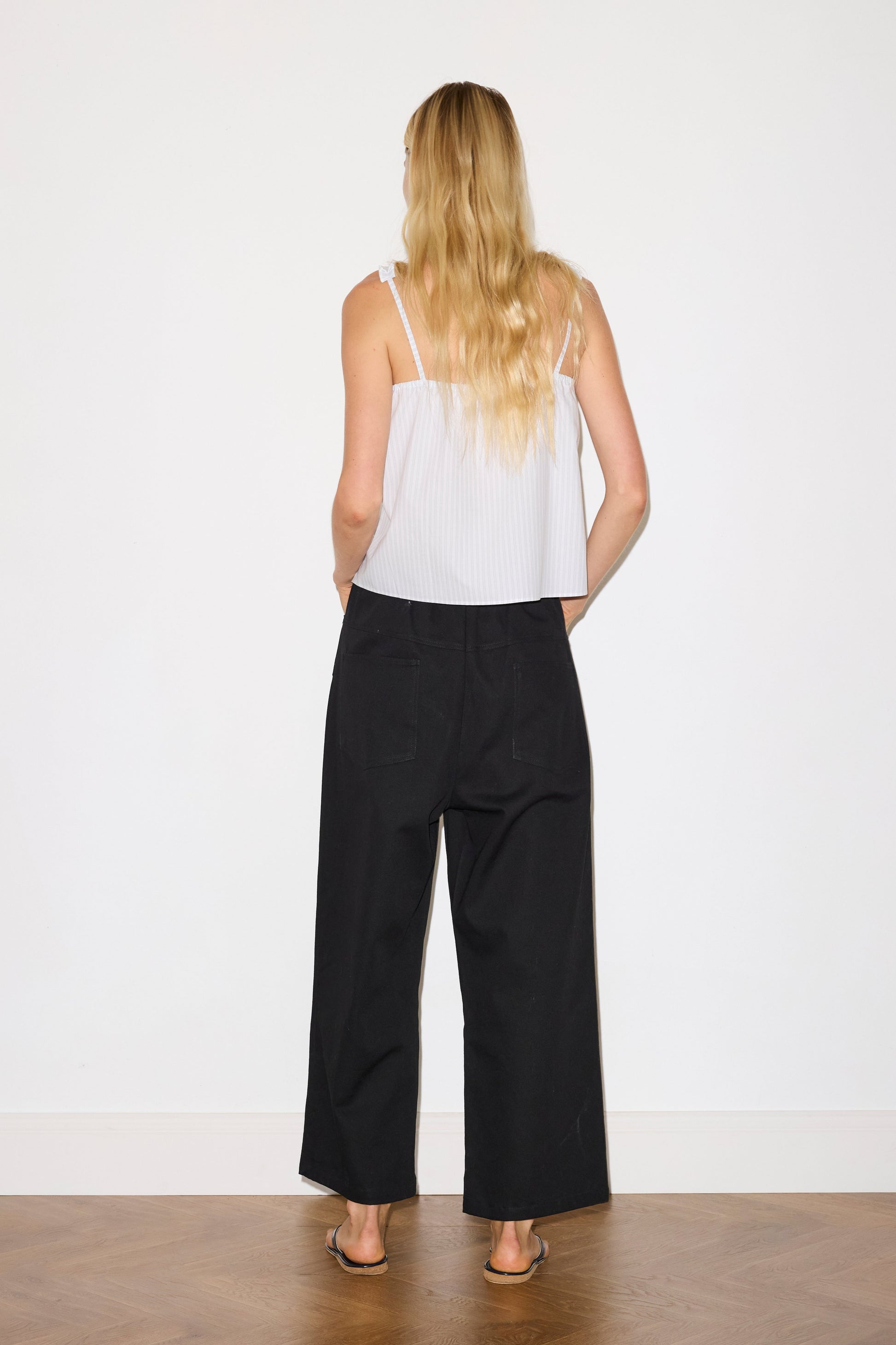 blonde female model wears a sage and white striped relaxed-fit top featuring self-fabric ruffles along the straps and center front, with a tie front detail. designed with elasticated gathering for ease and comfort, paired with a black Heavy- weighted relaxed-fit pant featuring a wide-leg silhouette, elasticated waistband, and square pocket detail