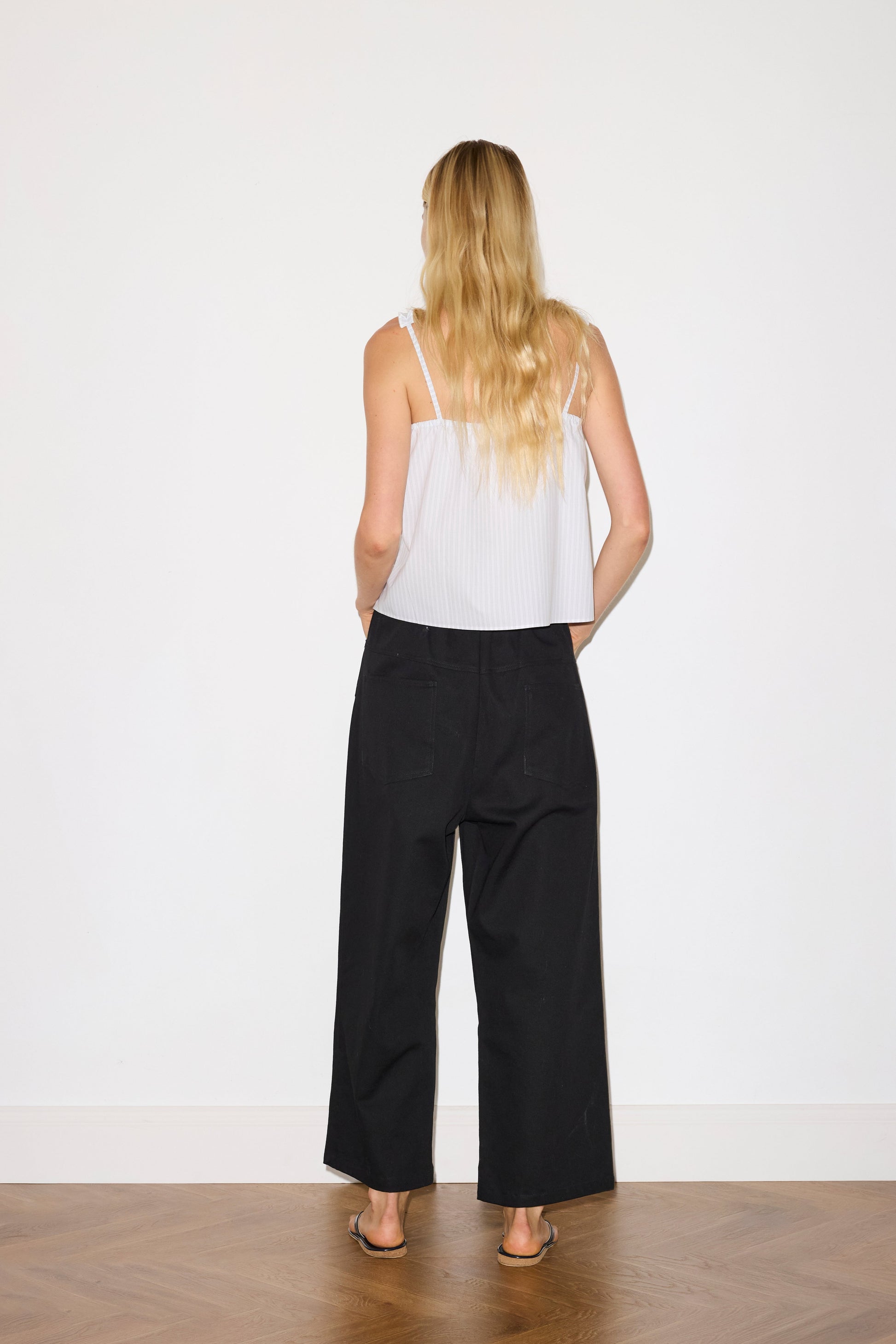 blonde haired female model wears a black Heavy- weighted relaxed-fit pant featuring a wide-leg silhouette, elasticated waistband, and square pocket detail, worn with a sage and white striped a relaxed-fit top featuring self-fabric ruffles along the straps and center front, with a tie front detail. designed with elasticated gathering for ease and comfort