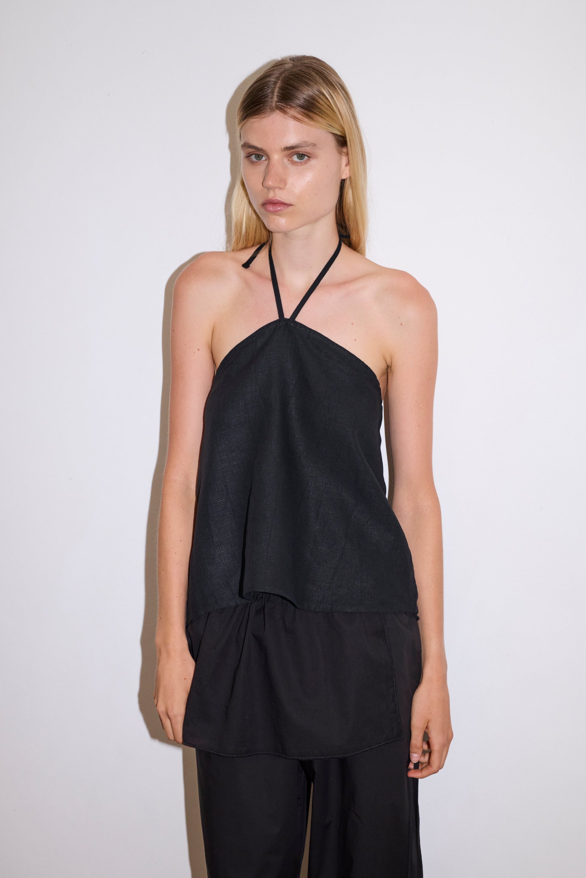 blonde haired female model wears a black relaxed halter top features flattering straight neckline, fine tie halter straps and snap closure at back with button detail, paired with a black ankle length pant features a soft minimal elastic waist with side seam pockets in a straight leg.