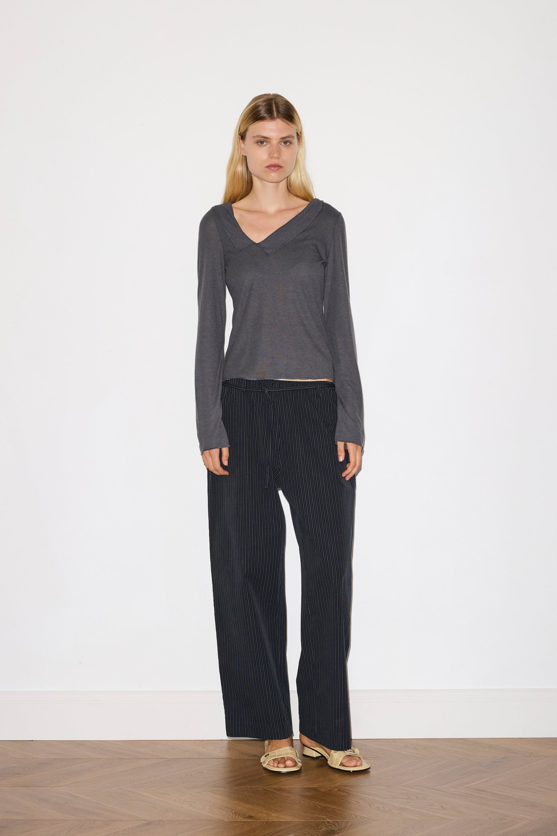 blonde female model wears a blue grey sheer fine knit long sleeved top with asymmetric collared neckline, featuring baby locked finishes and a relaxed fit, paired with a a full length, relaxed , mid-low waisted trouser with a removable string belt with mini silver buckle detail in yellow and navy stripe