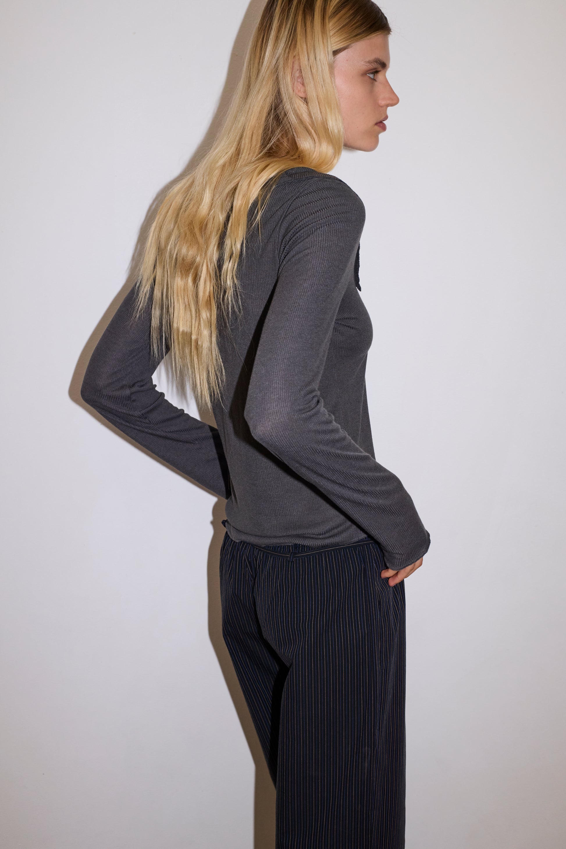 blonde female model wears a blue grey sheer fine knit long sleeved top with asymmetric collared neckline, featuring baby locked finishes and a relaxed fit, paired with a a full length, relaxed , mid-low waisted trouser with a removable string belt with mini silver buckle detail in yellow and navy stripe