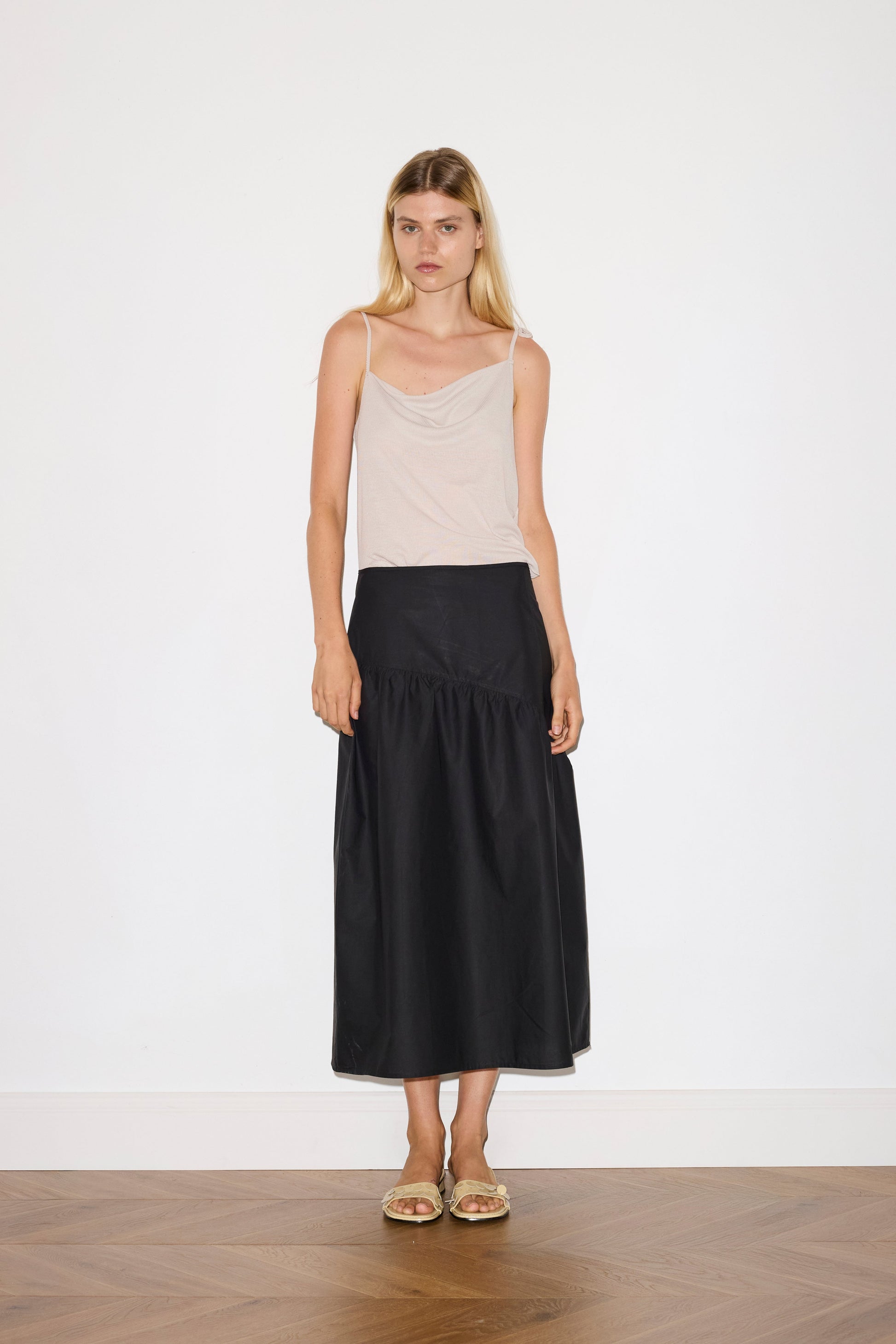 blonde female model wears a beige minimalist tank-style top featuring raw edge detailing and an asymmetrical tie strap design for a unique and adjustable fit. made with a lightweight ribbed texture, worn with a black ankle length, straight leg pant features a soft minimal elastic waist with front seam pockets and button detail