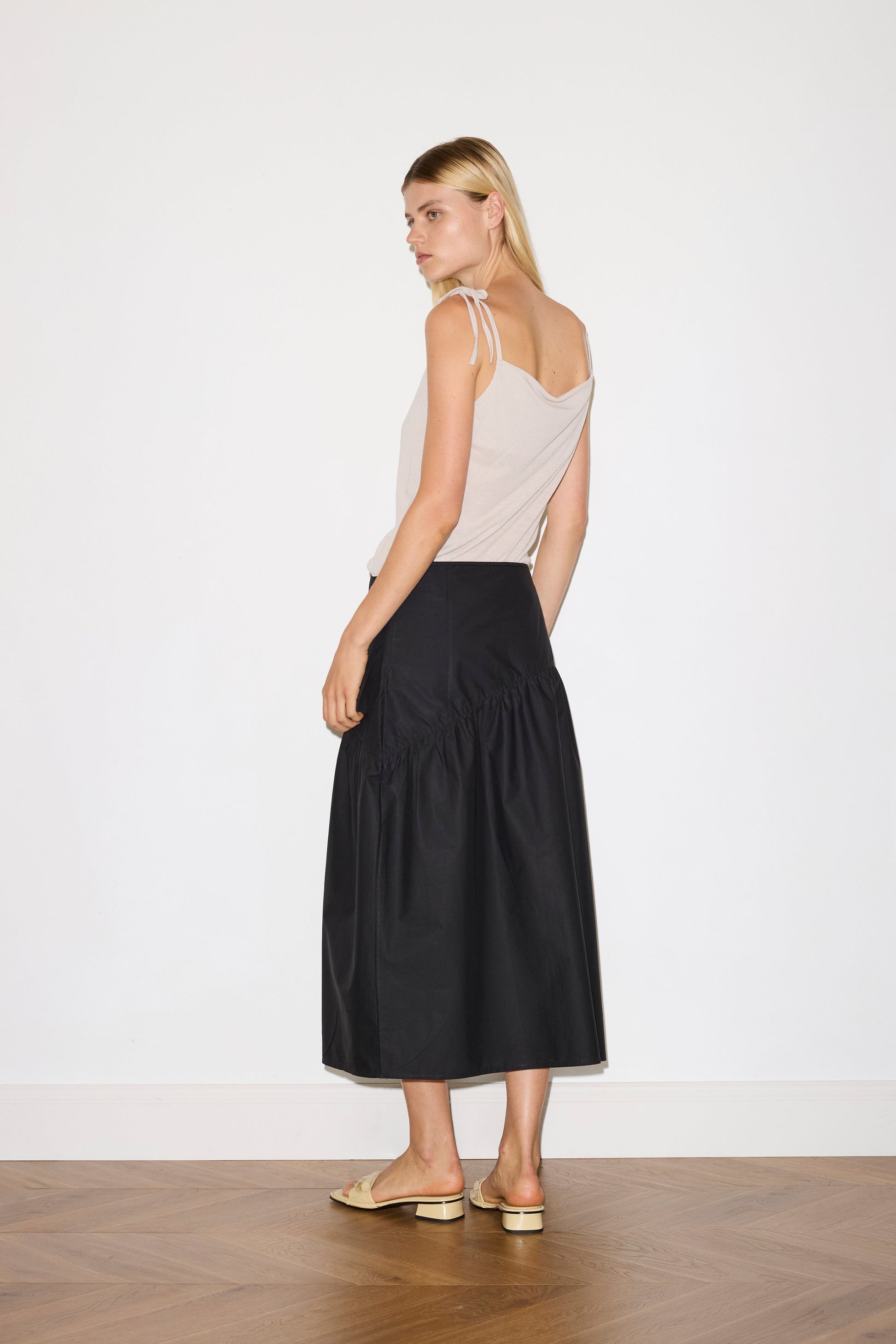 blonde female model wears a beige minimalist tank-style top featuring raw edge detailing and an asymmetrical tie strap design for a unique and adjustable fit. made with a lightweight ribbed texture, worn with a black ankle length, straight leg pant features a soft minimal elastic waist with front seam pockets and button detail