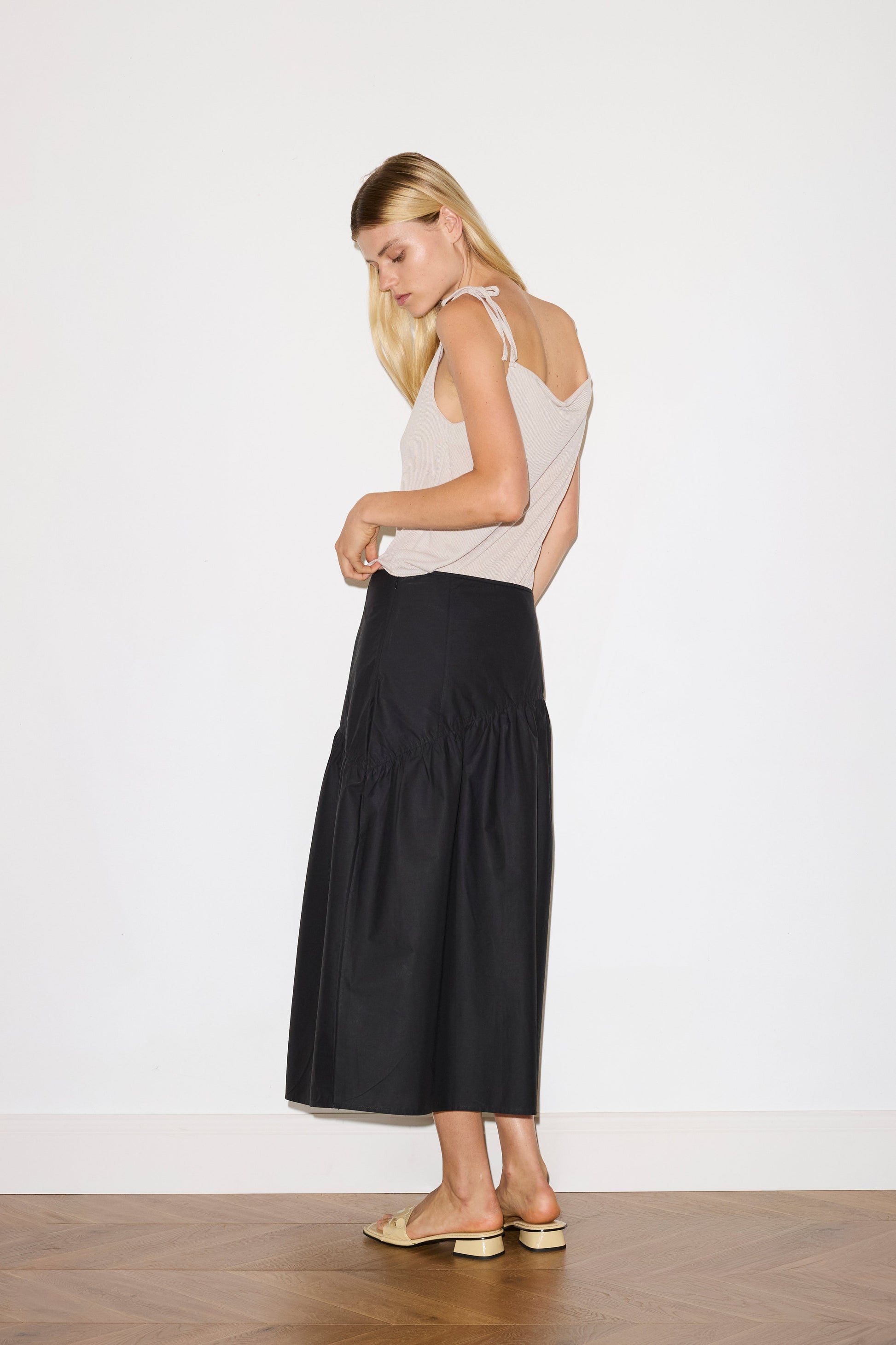 blonde female model wears a beige minimalist tank-style top featuring raw edge detailing and an asymmetrical tie strap design for a unique and adjustable fit. made with a lightweight ribbed texture, worn with a black ankle length, straight leg pant features a soft minimal elastic waist with front seam pockets and button detail