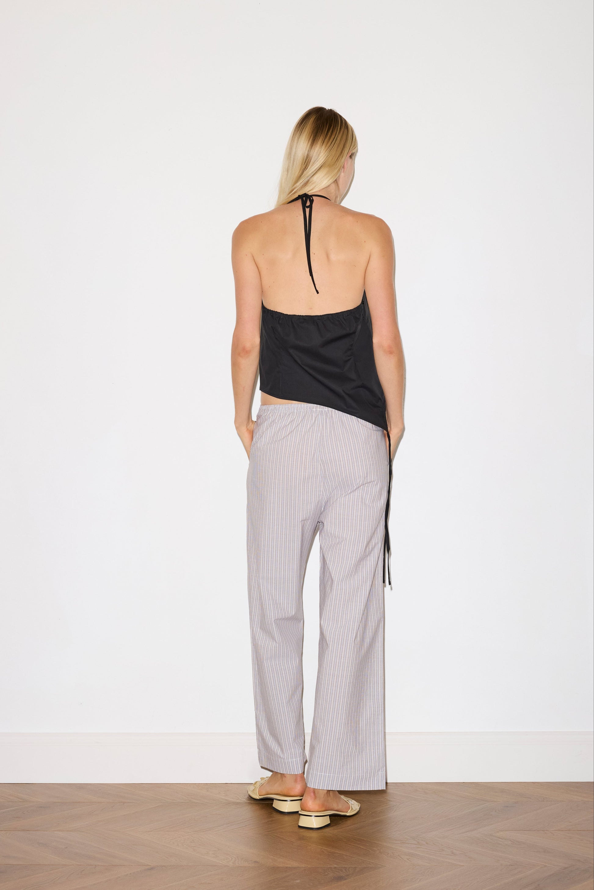 blonde female model wears a black relaxed halter top features flattering curved neckline, assymetrical bottom hem feature with side tie opening, keyhole with button closure at side and fine tie halter straps, paired with a lilac, blue and white striped organic cotton trouser with an elasticated waist