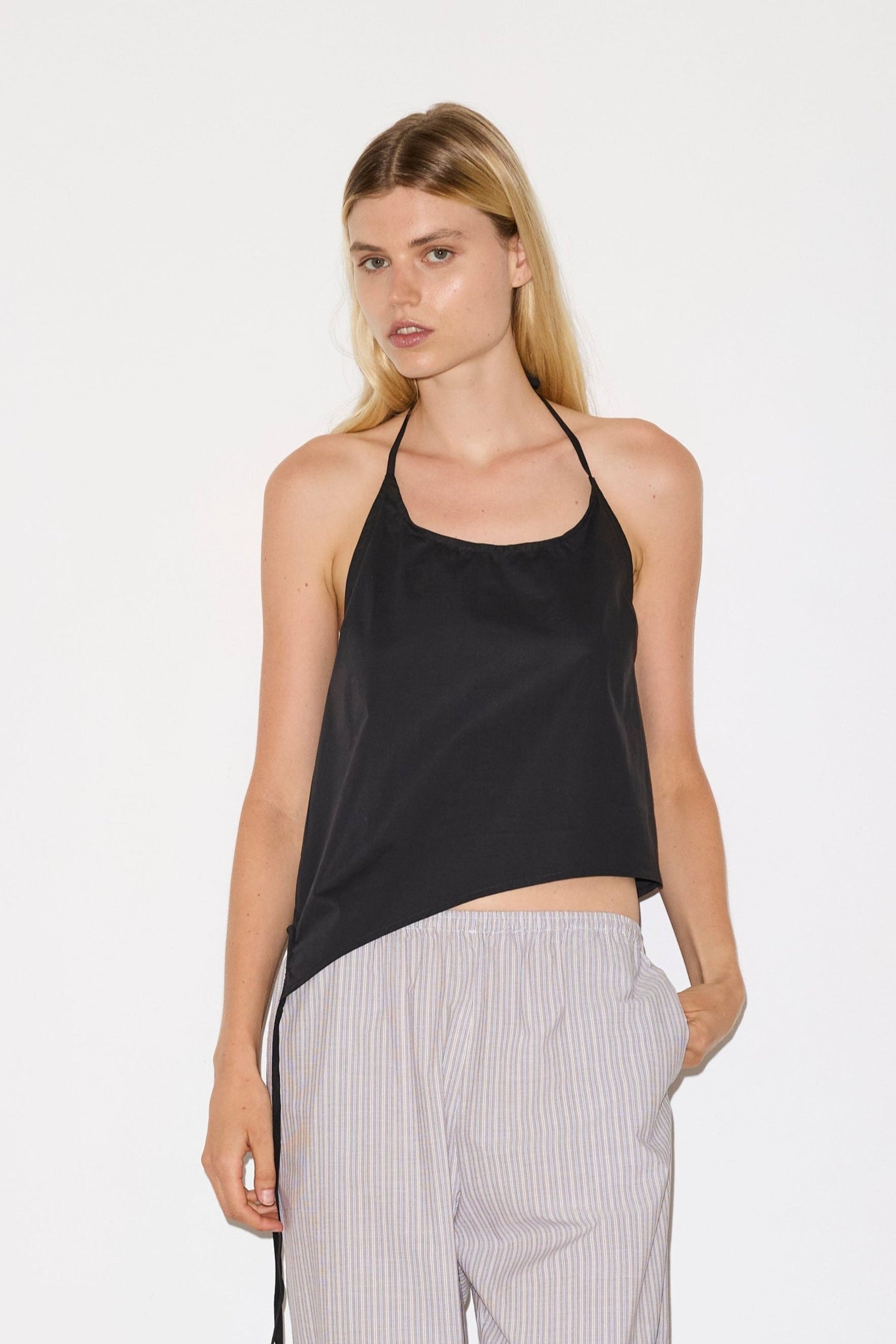 blonde female model wears a black relaxed halter top features flattering curved neckline, assymetrical bottom hem feature with side tie opening, keyhole with button closure at side and fine tie halter straps, paired with a lilac, blue and white striped organic cotton trouser with an elasticated waist