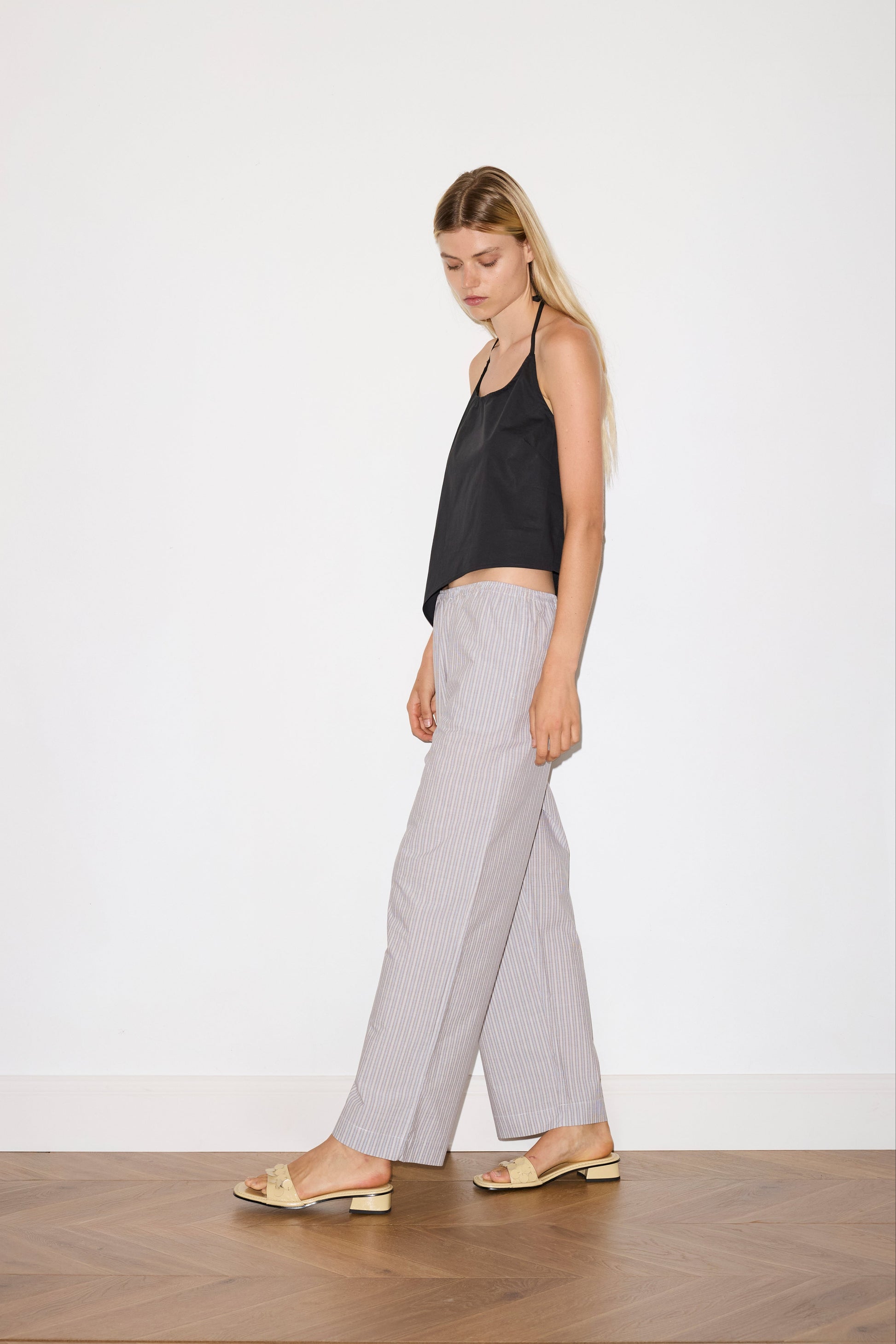 blonde female model wears a black relaxed halter top features flattering curved neckline, assymetrical bottom hem feature with side tie opening, keyhole with button closure at side and fine tie halter straps, paired with a lilac, blue and white striped organic cotton trouser with an elasticated waist