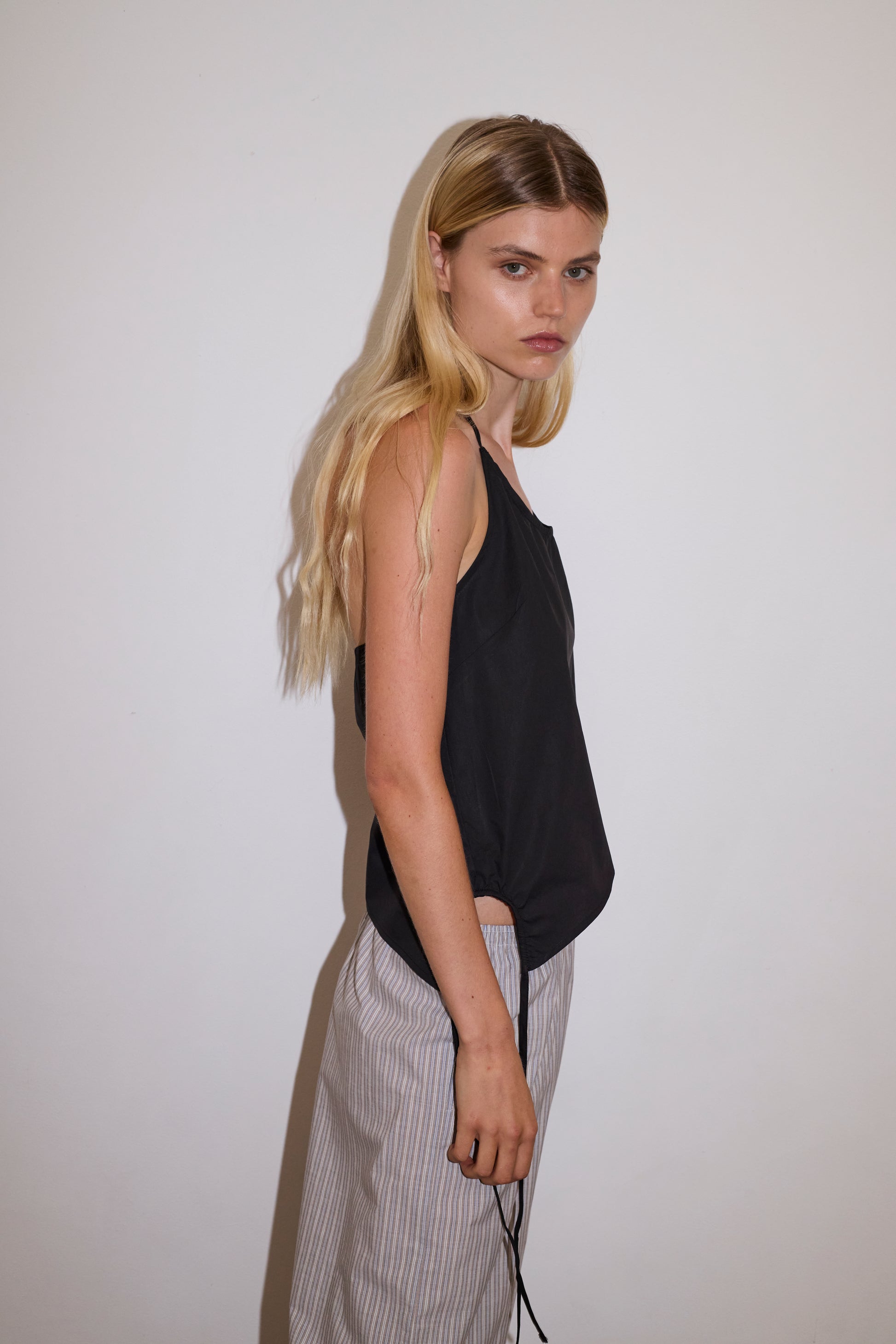 blonde female model wears a black relaxed halter top features flattering curved neckline, assymetrical bottom hem feature with side tie opening, keyhole with button closure at side and fine tie halter straps, paired with a lilac, blue and white striped organic cotton trouser with an elasticated waist