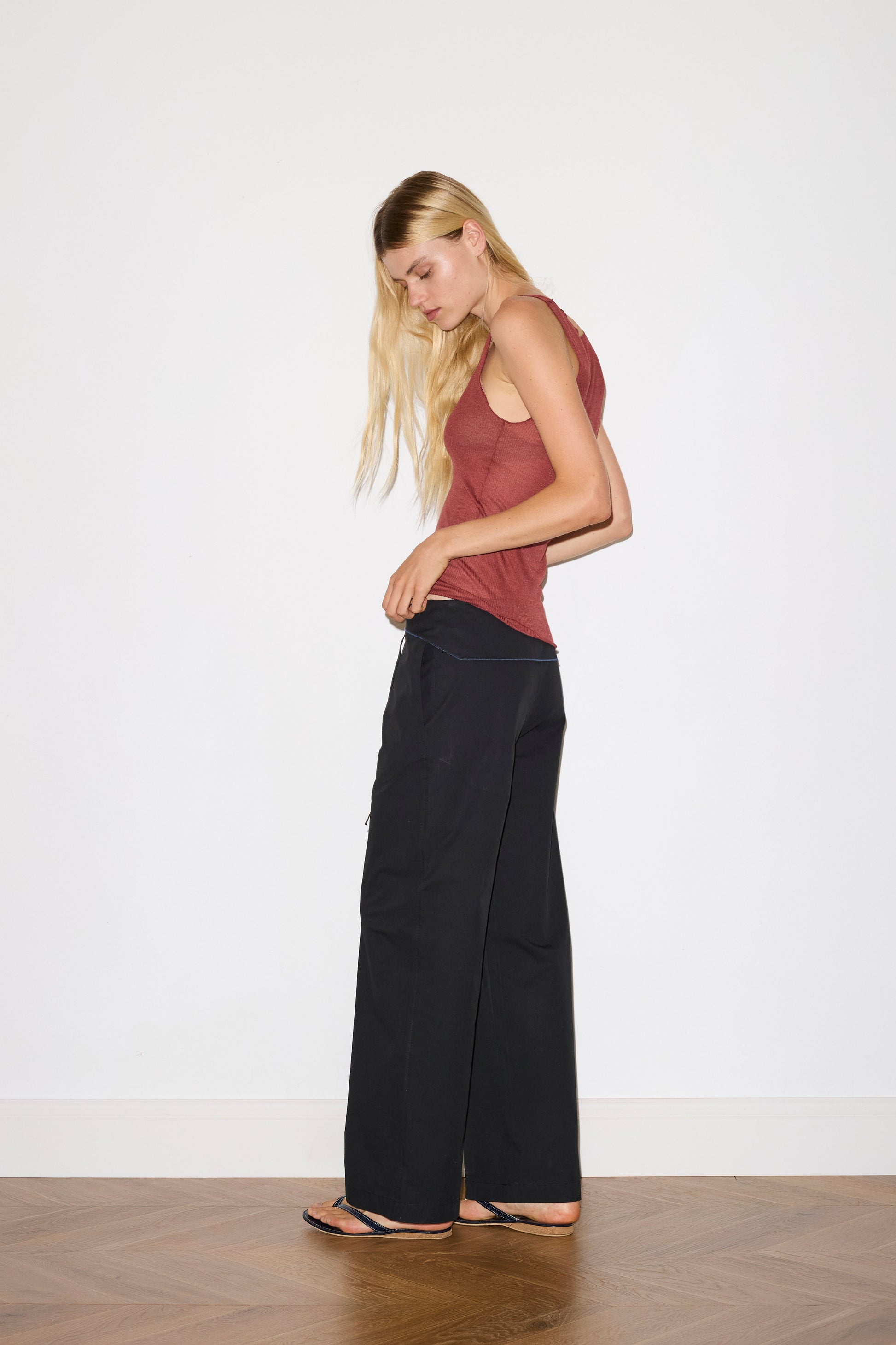 blonde female model wears a knit tank in crimson red in a sheer material, worn with a full-length black trouser with tie belt detail and size zip closure.