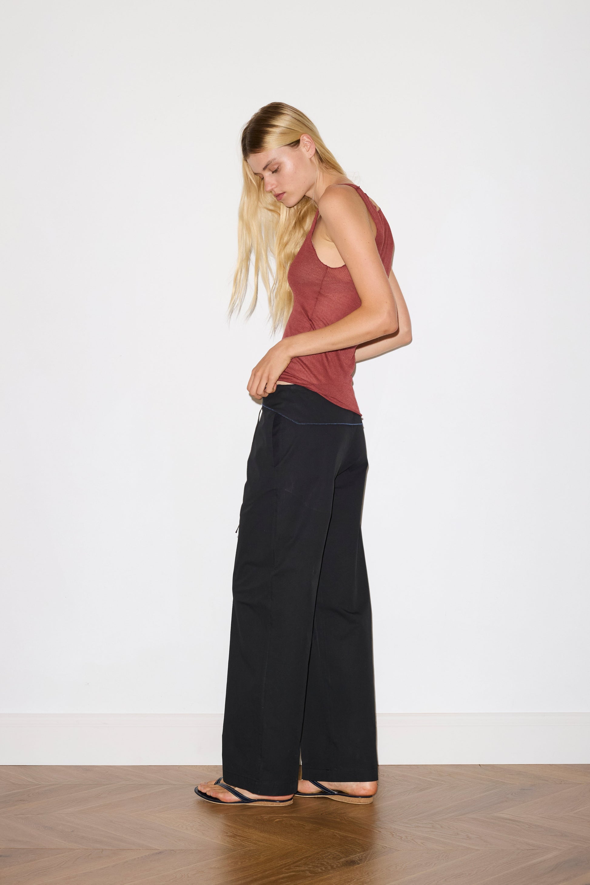 blonde female model wears a knit tank in crimson red in a sheer material, worn with a full-length black trouser with tie belt detail and size zip closure.