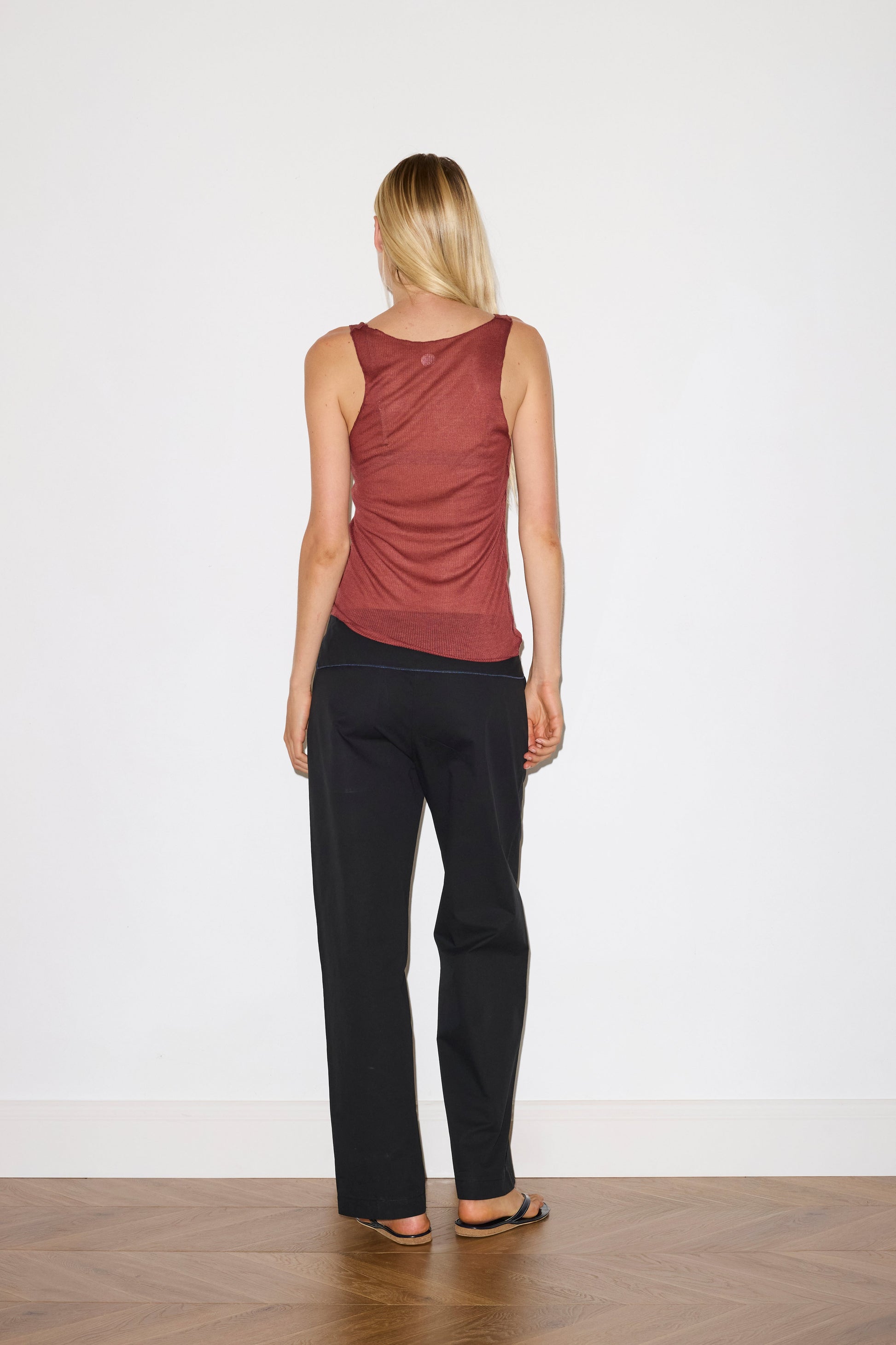 blonde female model wears a knit tank in crimson red in a sheer material, worn with a full-length black trouser with tie belt detail and size zip closure.