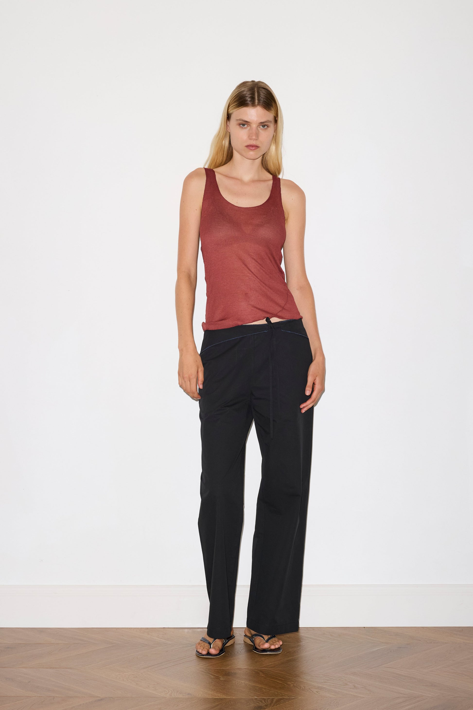 blonde female model wears a knit tank in crimson red in a sheer material, worn with a full-length black trouser with tie belt detail and size zip closure.