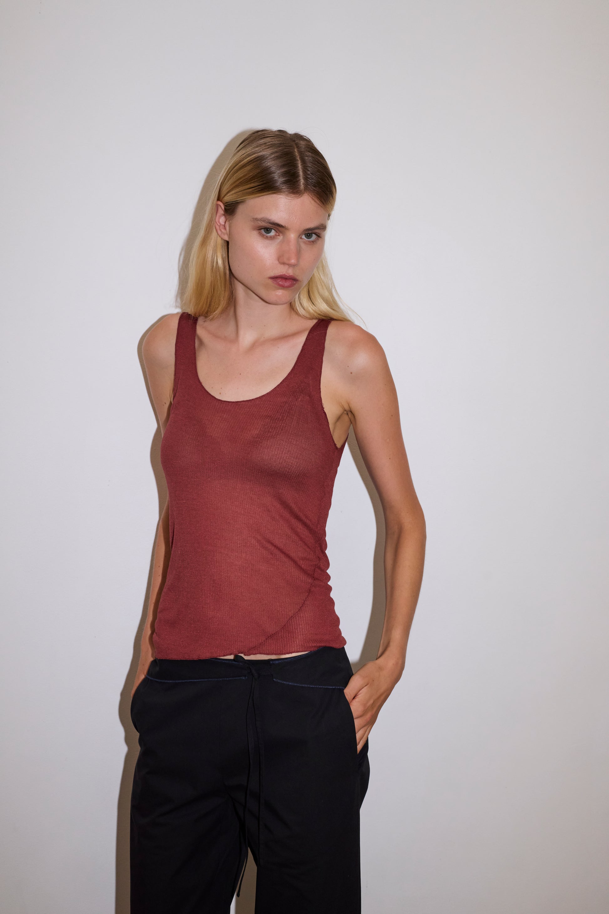 blonde female model wears a knit tank in crimson red in a sheer material, worn with a full-length black trouser with tie belt detail and size zip closure.