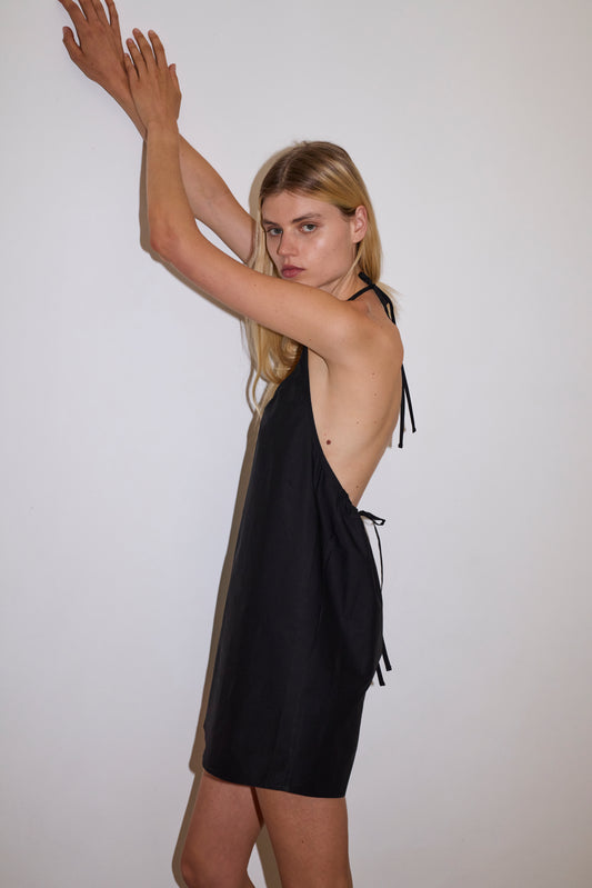 blonde female model wears a black short-length, halter dress featuring a rounded v neckline with adjustable ties around the neck. designed with external center-front tucks created by an internal elastic. the back neckline includes a keyhole detail with functional ties