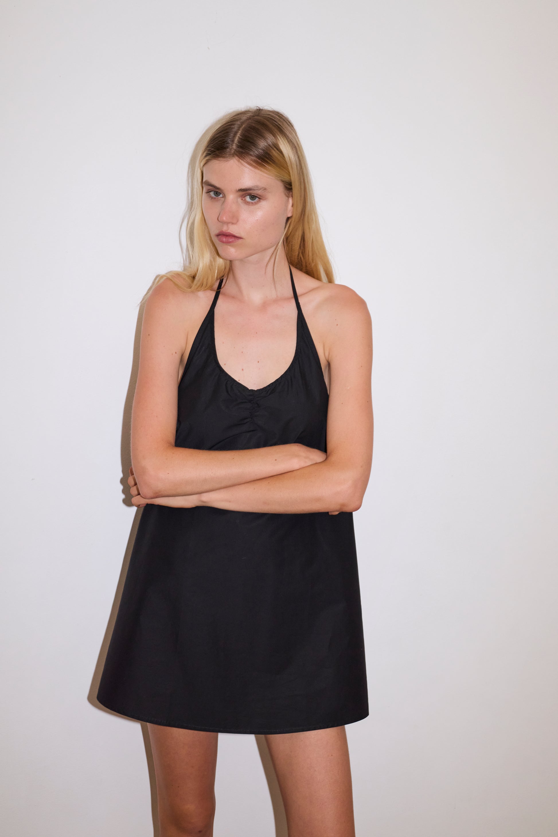 blonde female model wears a black short-length, halter dress featuring a rounded v neckline with adjustable ties around the neck. designed with external center-front tucks created by an internal elastic. the back neckline includes a keyhole detail with functional ties