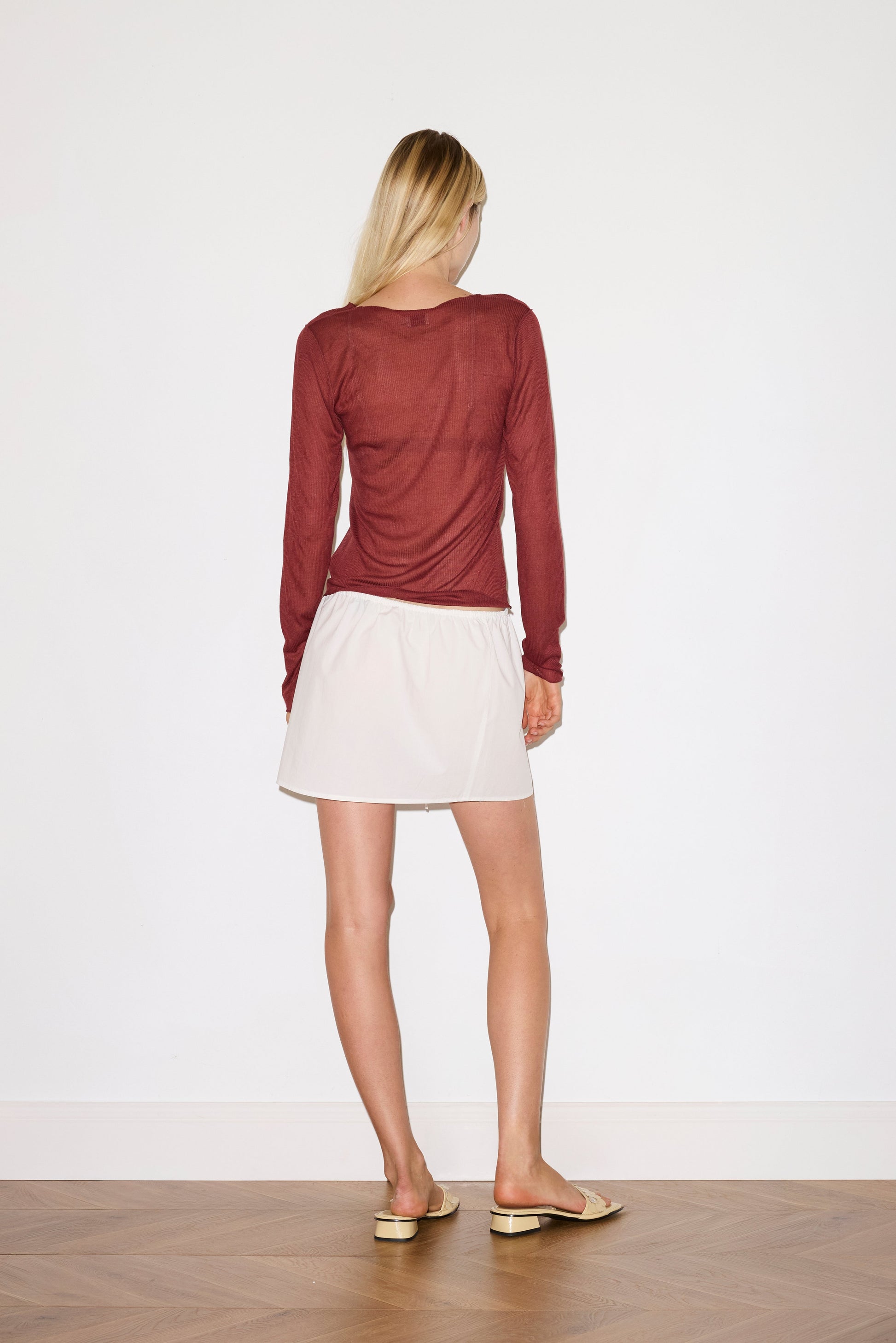 blonde female models wears a red a sheer fine knit long sleeved top with exposed seams and tag, featuring baby locked finishes and a relaxed fit, worn with a white simple mini skirt designed to sit at low waist features adjustable tie and ruffle edge on side with button detail