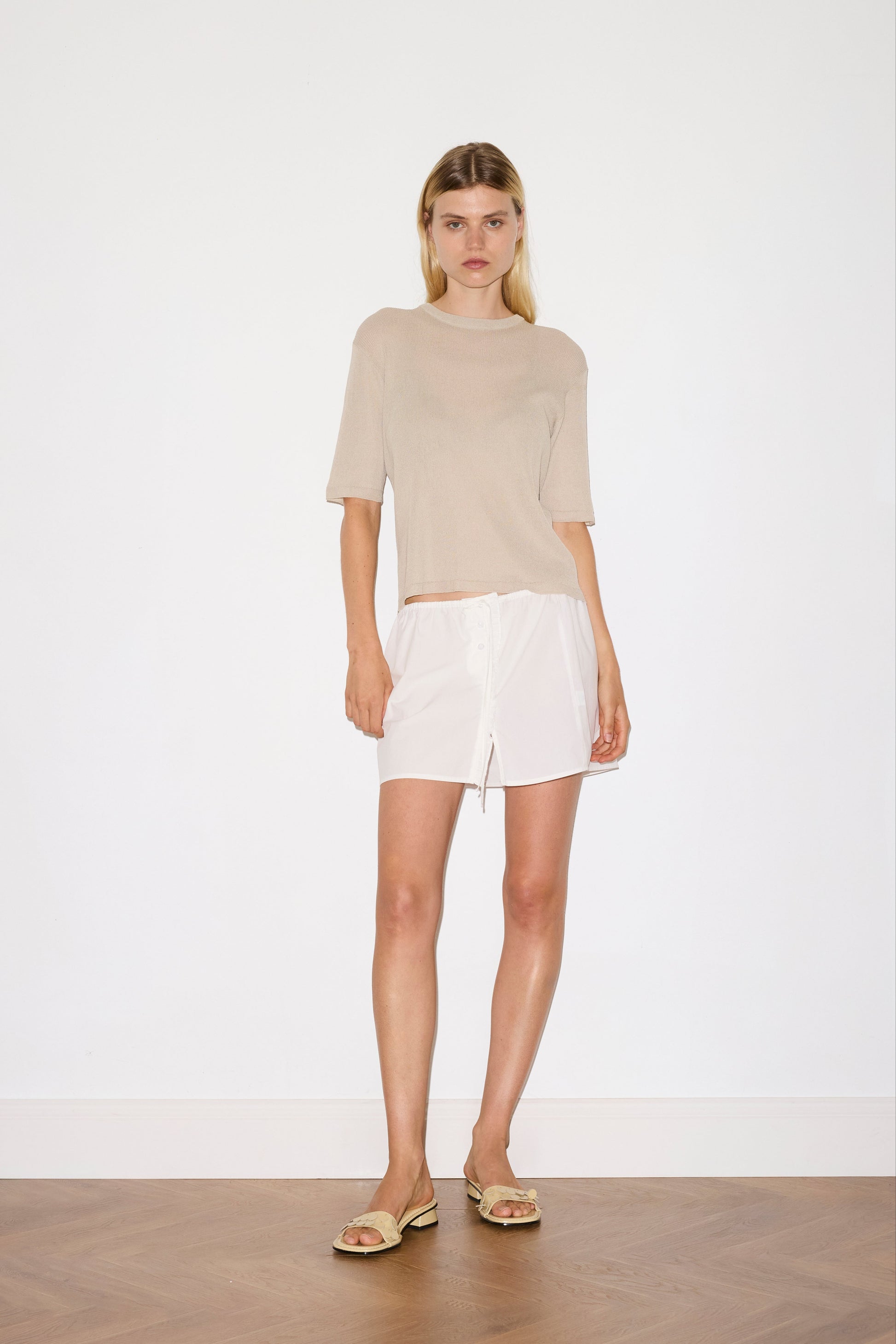 blonde female model wears a beige fully fashioned, relaxed short-sleeve tee featuring a ribbed texture and a split back detail with flat tie closures, worn with a white simple mini skirt designed to sit at low waist features adjustable tie and ruffle edge on side with button detail