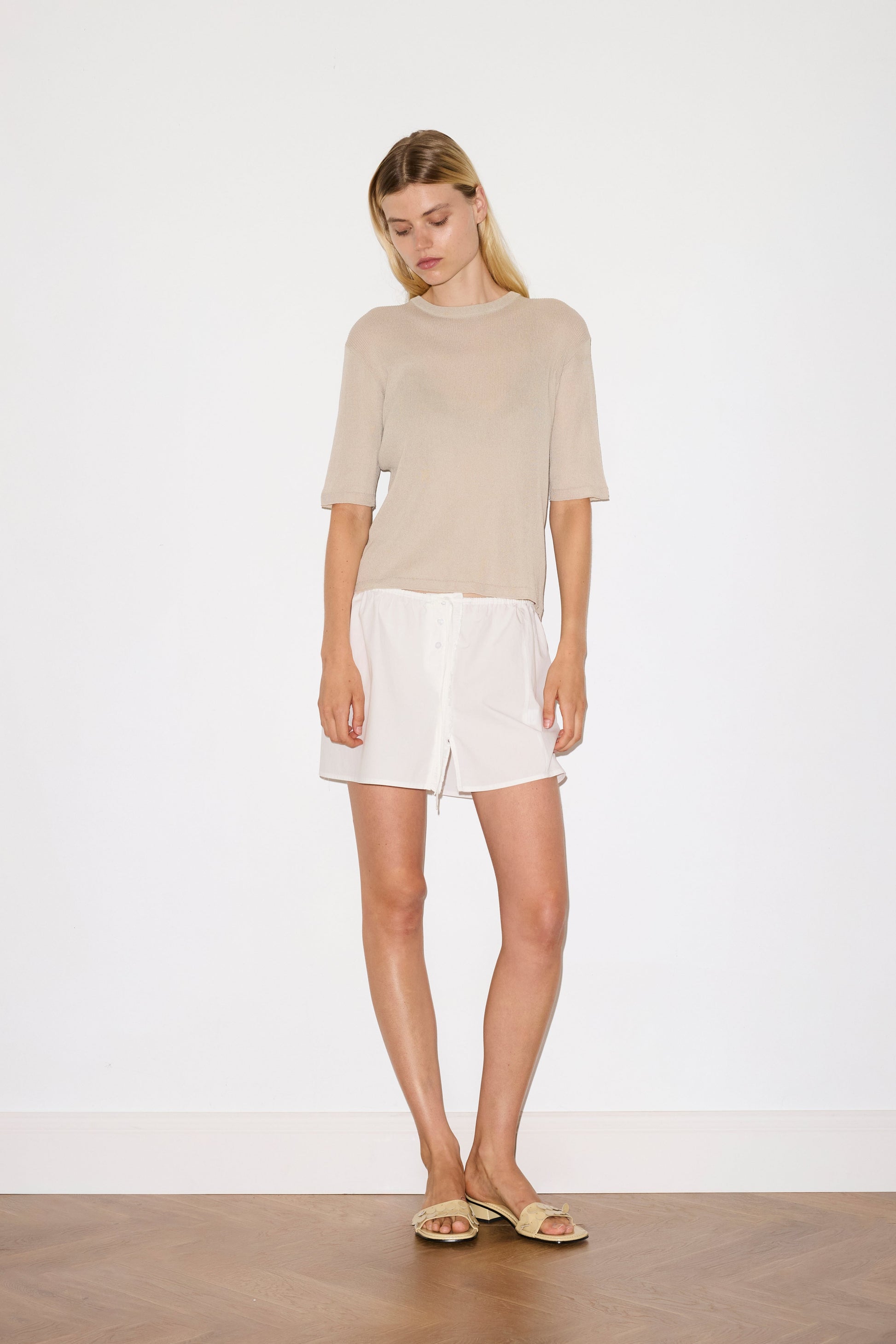 blonde female model wears a beige fully fashioned, relaxed short-sleeve tee featuring a ribbed texture and a split back detail with flat tie closures, worn with a white simple mini skirt designed to sit at low waist features adjustable tie and ruffle edge on side with button detail