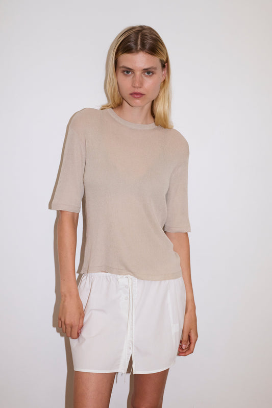 blonde female model wears a beige fully fashioned, relaxed short-sleeve tee featuring a ribbed texture and a split back detail with flat tie closures, worn with a white simple mini skirt designed to sit at low waist features adjustable tie and ruffle edge on side with button detail