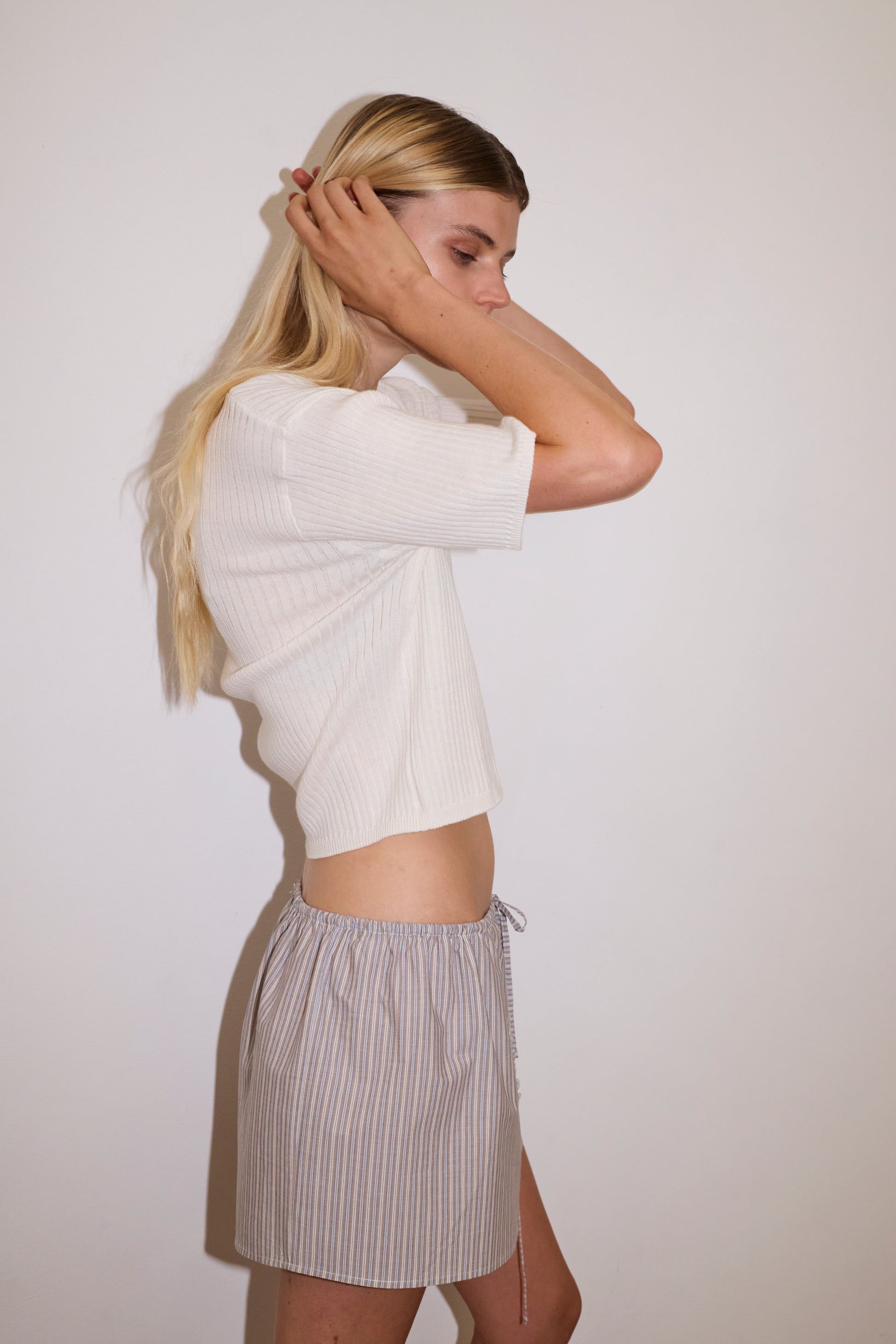 blonde female model wears a white fully fashioned ribbed knit features a flattering deep v neckline, mid length sleeve and button front design feature in a relaxed fit, worn with a lilac and blue simple mini skirt designed to sit at low waist features adjustable tie and ruffle edge on side with button detail