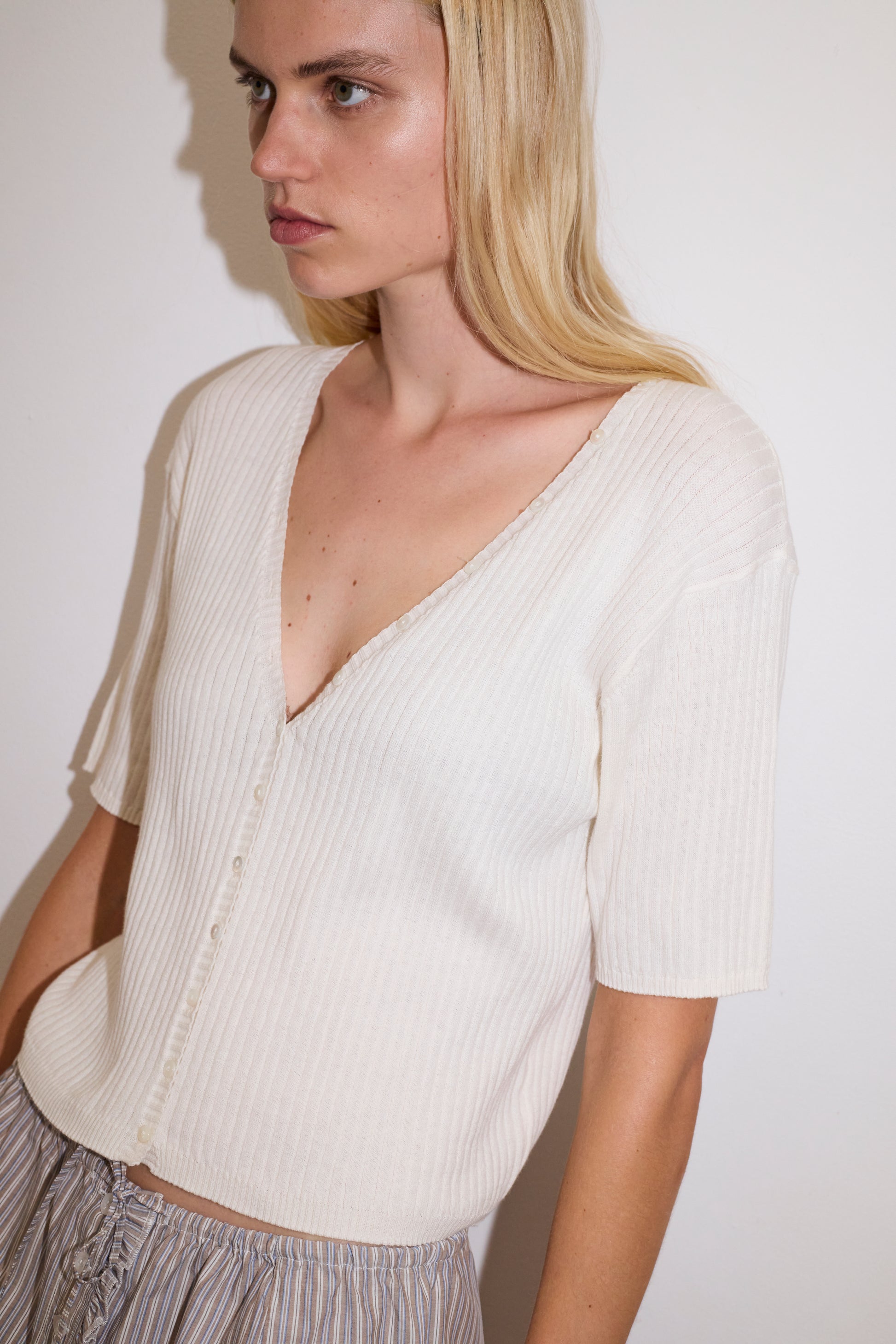 blonde female model wears a white fully fashioned ribbed knit features a flattering deep v neckline, mid length sleeve and button front design feature in a relaxed fit, worn with a lilac and blue simple mini skirt designed to sit at low waist features adjustable tie and ruffle edge on side with button detail