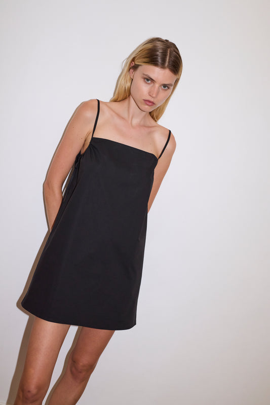 female blonde model wears a black lightweight, straight cut short length dress featuring gathering at the underarms with keyhole and ties on the right side