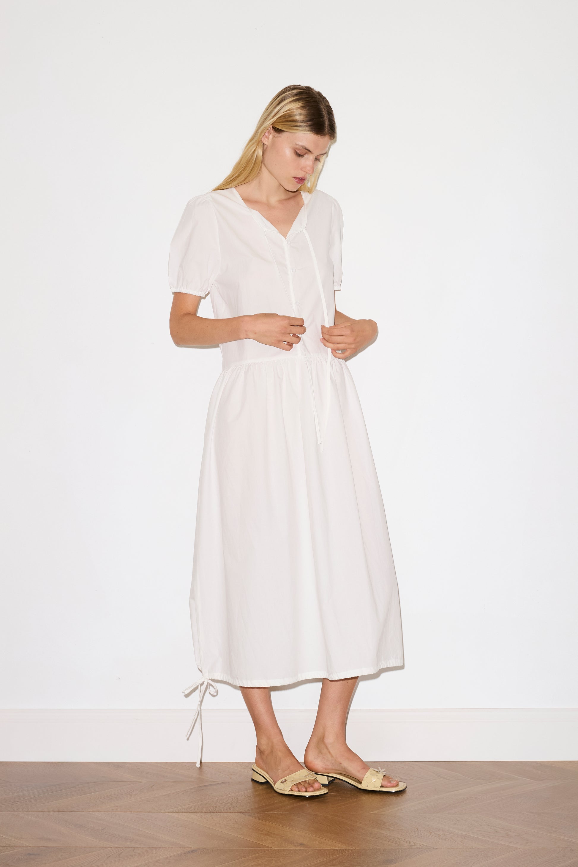 blonde female model wears a white elevated everyday dress in mid length features a flattering v neckline with tie, button front detail with a drop waist, and soft elastic blouson sleeve