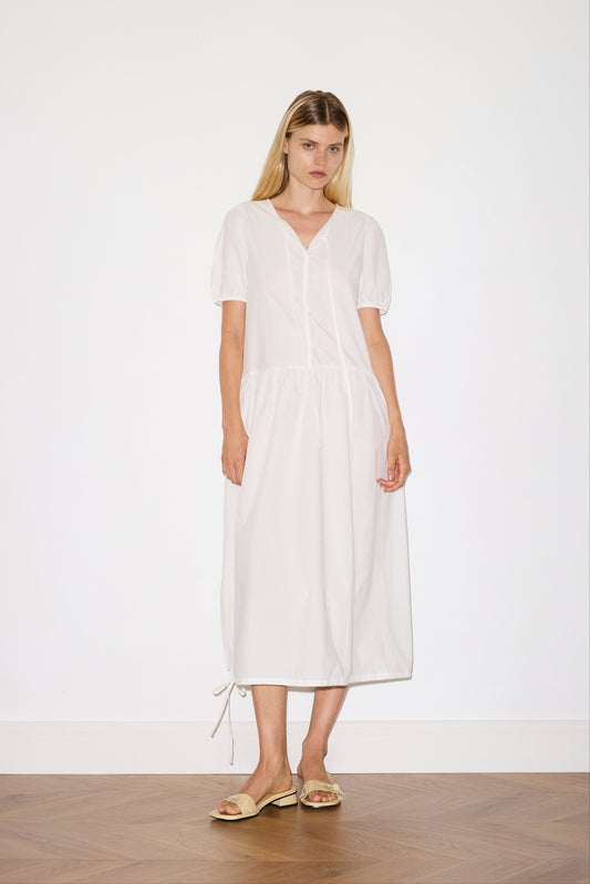 blonde female model wears a white elevated everyday dress in mid length features a flattering v neckline with tie, button front detail with a drop waist, and soft elastic blouson sleeve