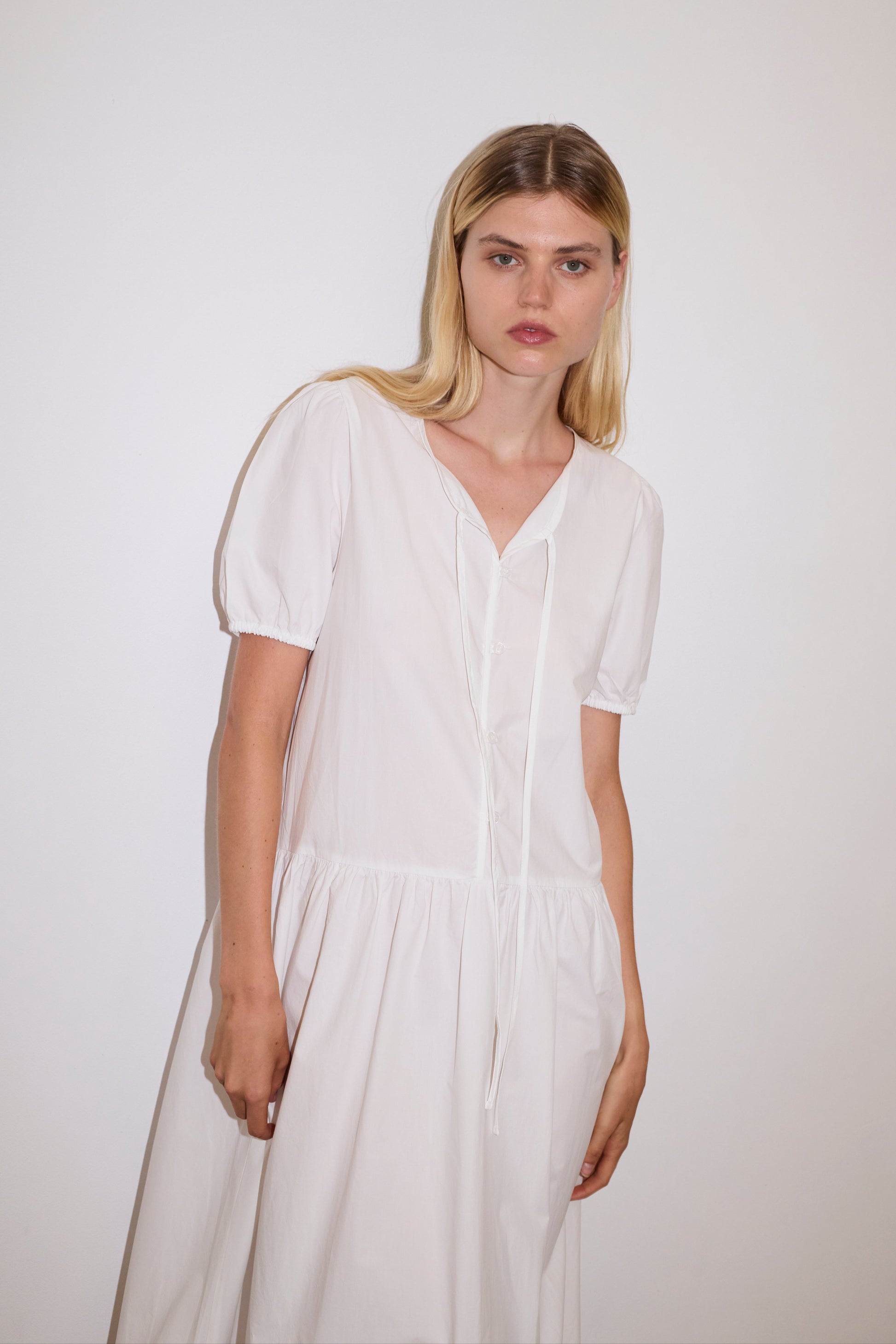 blonde female model wears a white elevated everyday dress in mid length features a flattering v neckline with tie, button front detail with a drop waist, and soft elastic blouson sleeve