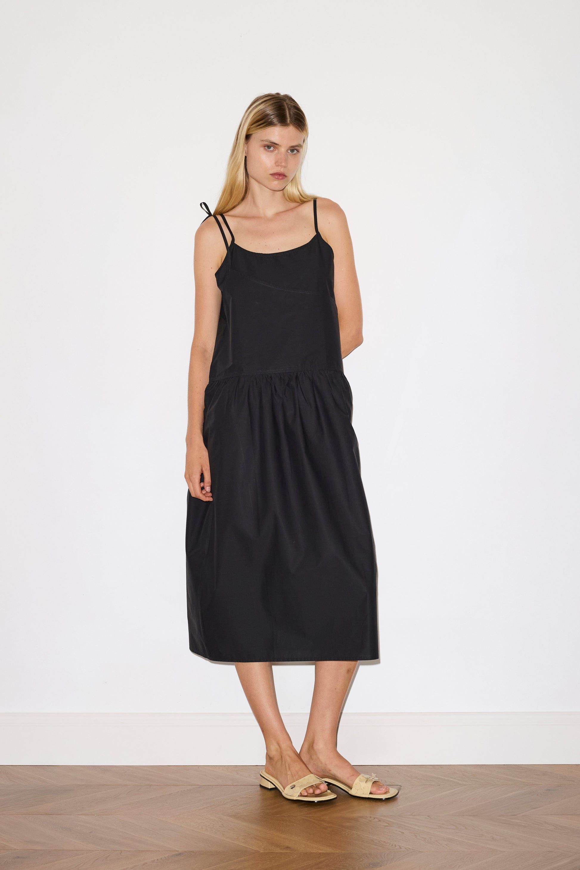 blonde female model wears a black mid-length dress featuring adjustable underlayer straps with sliders for a customised fit and a tied overlayer strap detail at the shoulder