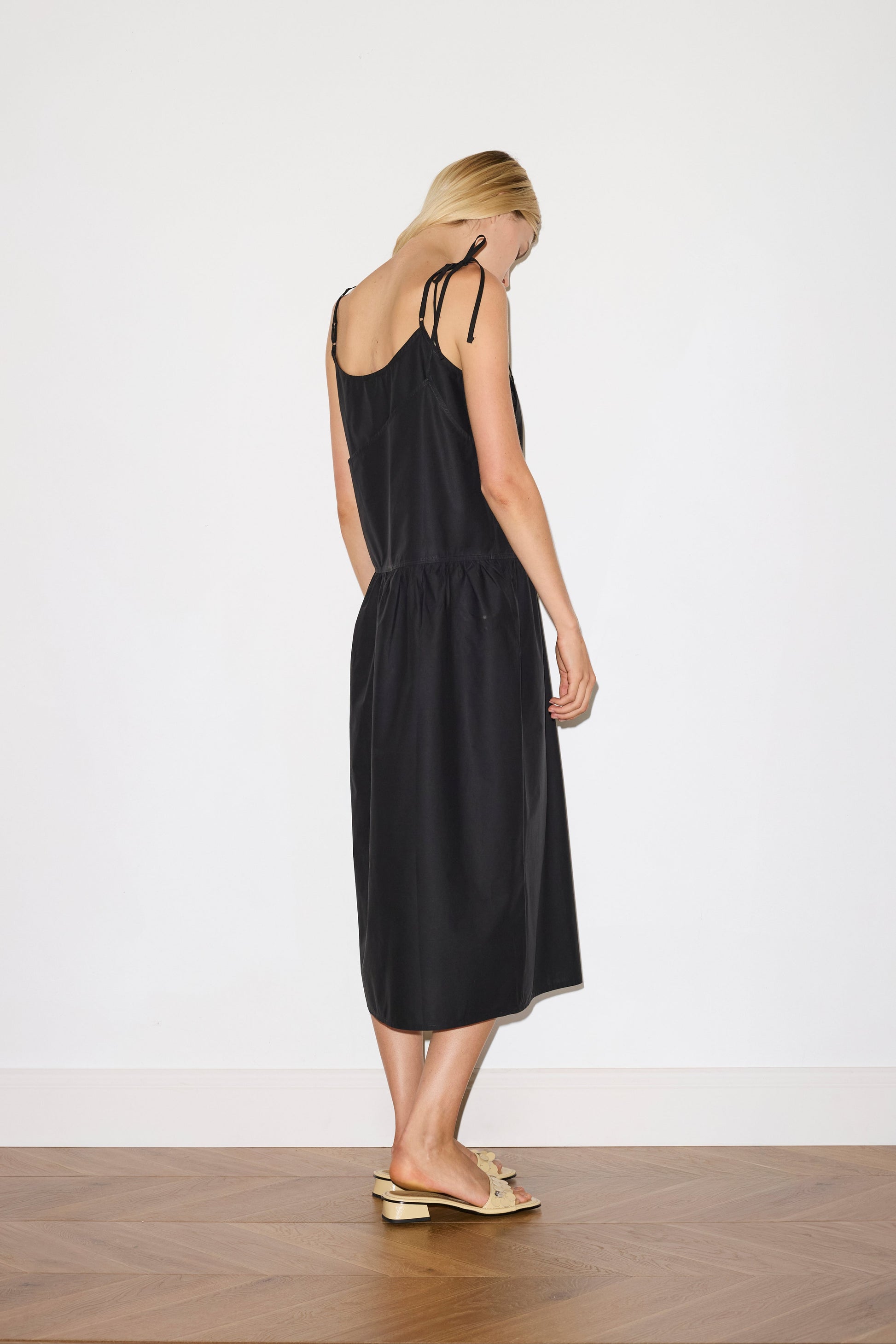 blonde female model wears a black mid-length dress featuring adjustable underlayer straps with sliders for a customised fit and a tied overlayer strap detail at the shoulder